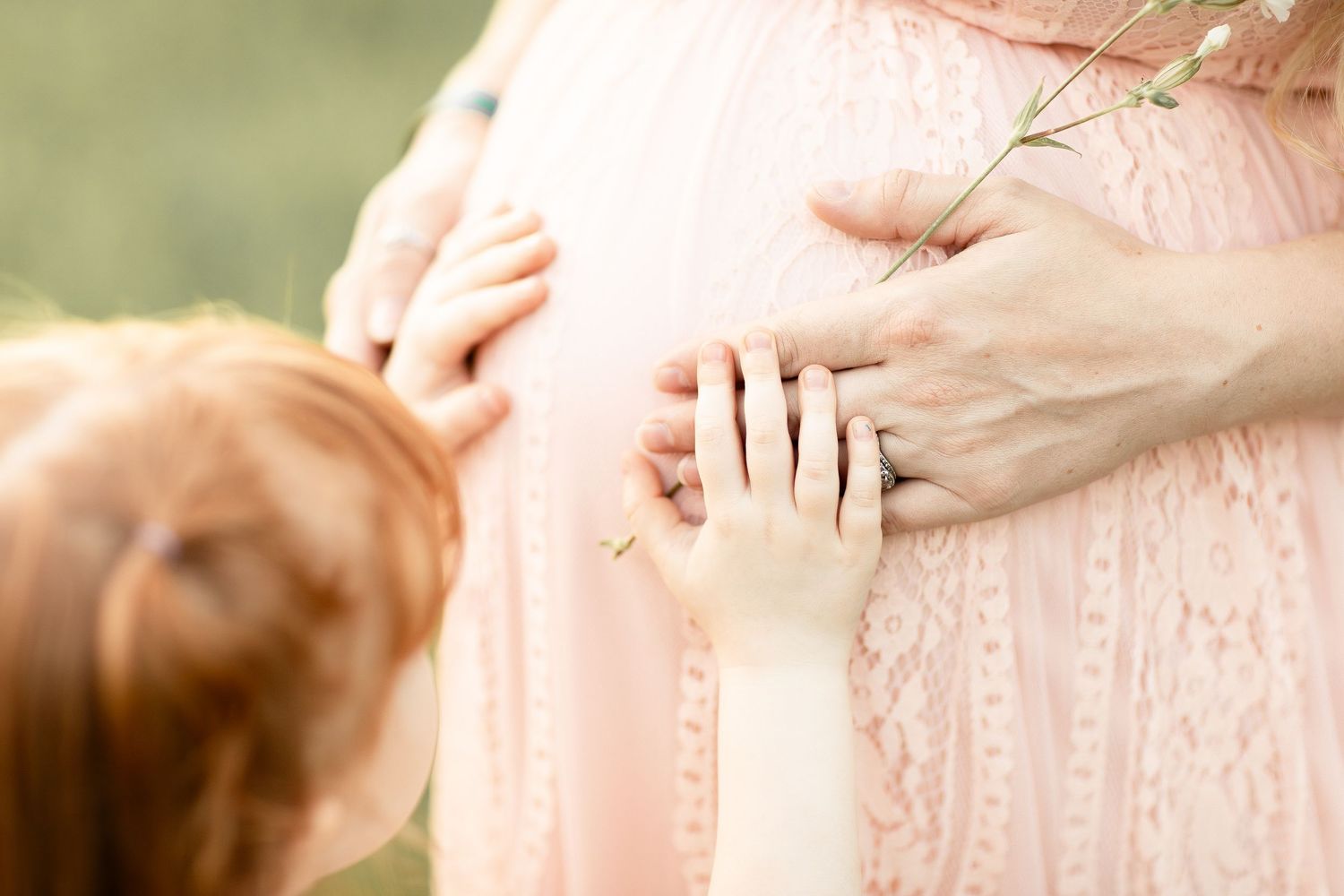 Close-up of mother and child hands gently touching pregnant belly in pink lace dress.