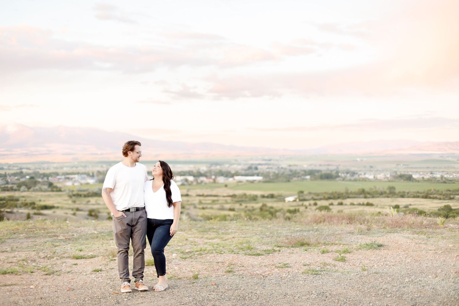A couple holding hands and gazing at each other on a hilltop with a scenic valley view.