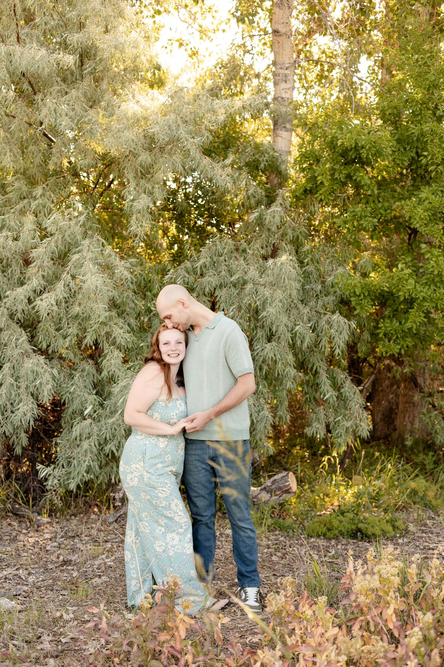 Couple embracing and kissing in a sunlit outdoor setting surrounded by green trees.