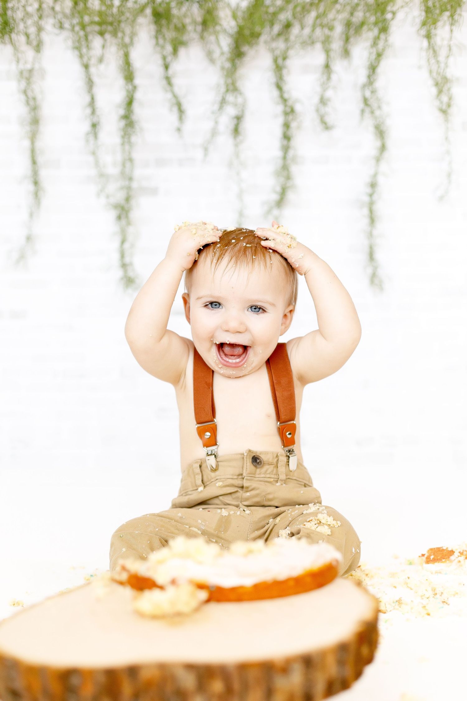 A joyful baby boy laughing with hands on head during a cake smash first birthday photoshoot.