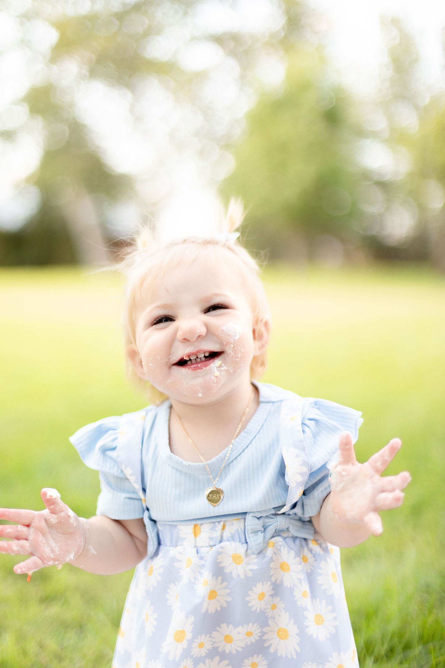 Happy baby girl with cake on face laughing in blue daisy dress during first birthday celebration.