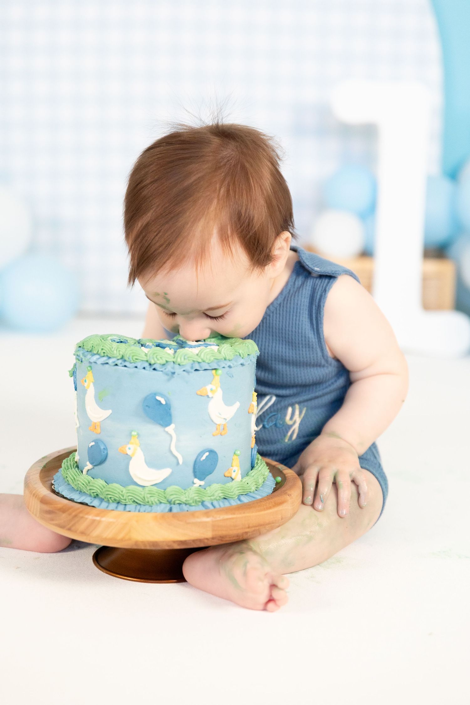 Baby boy leans in to taste blue duck-decorated first birthday cake on wooden cake stand.