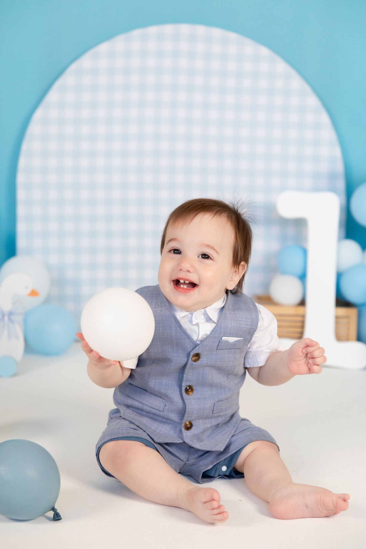 Happy baby boy sitting and holding a white balloon at a first birthday party with blue decor.