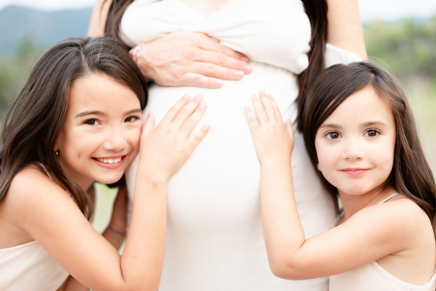Two young sisters smiling while hugging pregnant mothers baby bump outdoors.