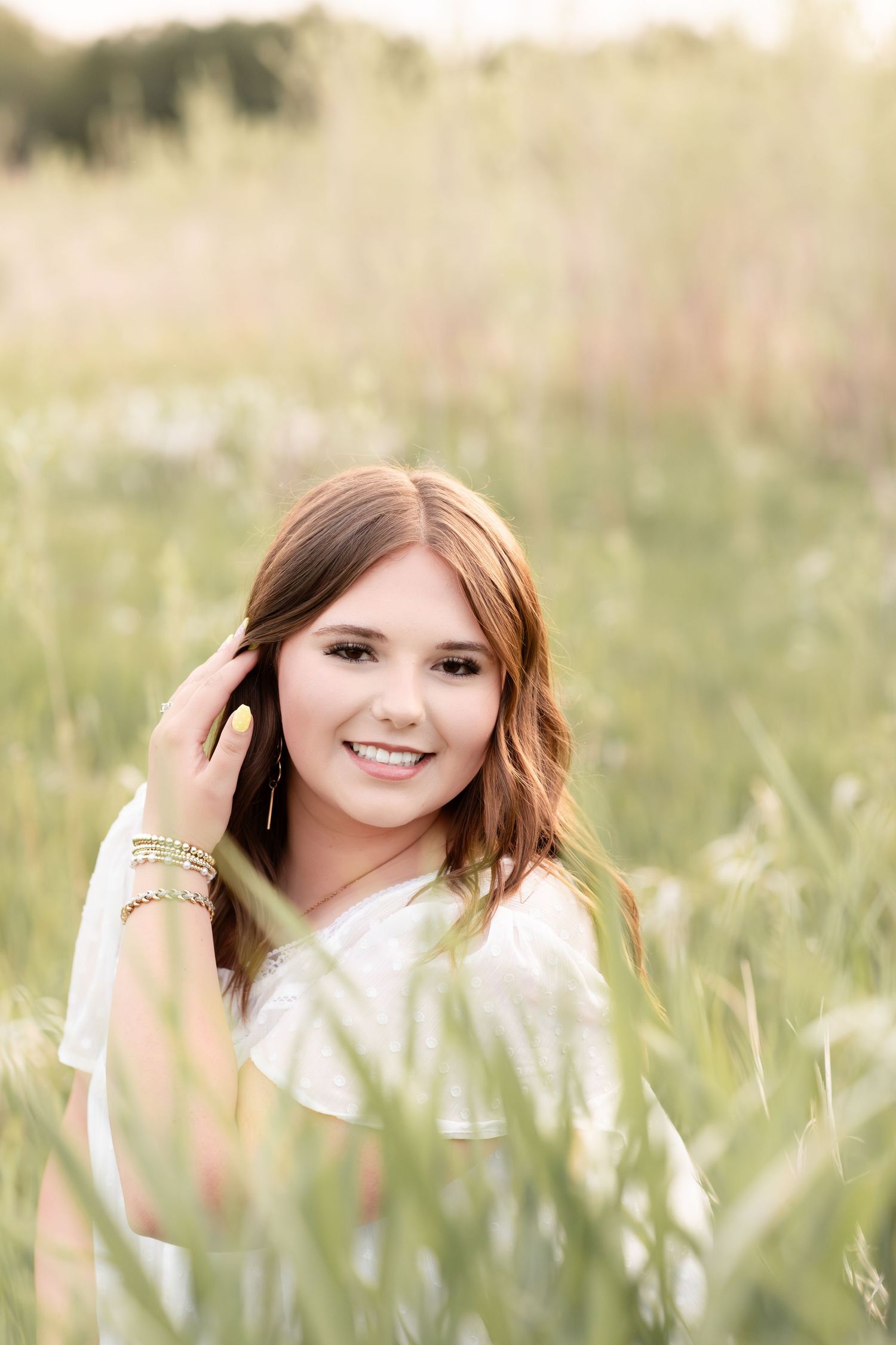 Smiling teenage girl with bracelet sitting in tall green grass field during golden hour.