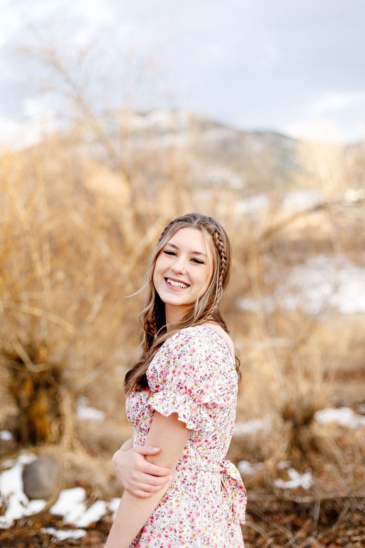 Smiling teenage girl in floral dress twirling among golden winter brush with snowy mountains.