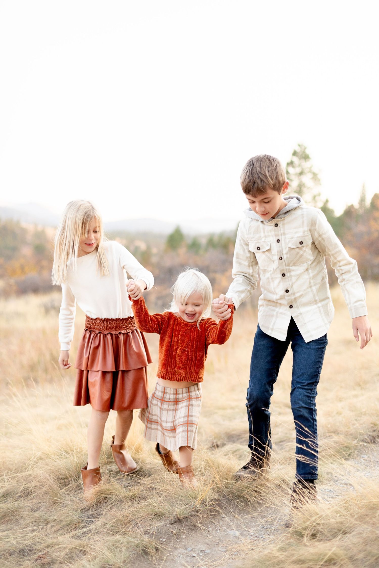 Older siblings swinging their young toddler sister by the hands along a dry grass field path.