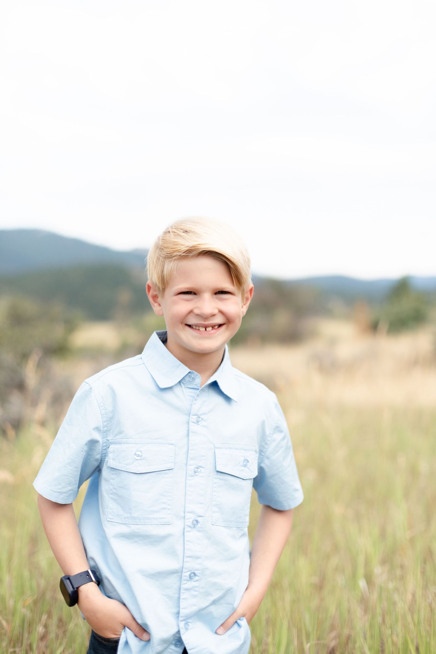 Smiling blonde boy in a light blue shirt standing in a grassy mountain field.