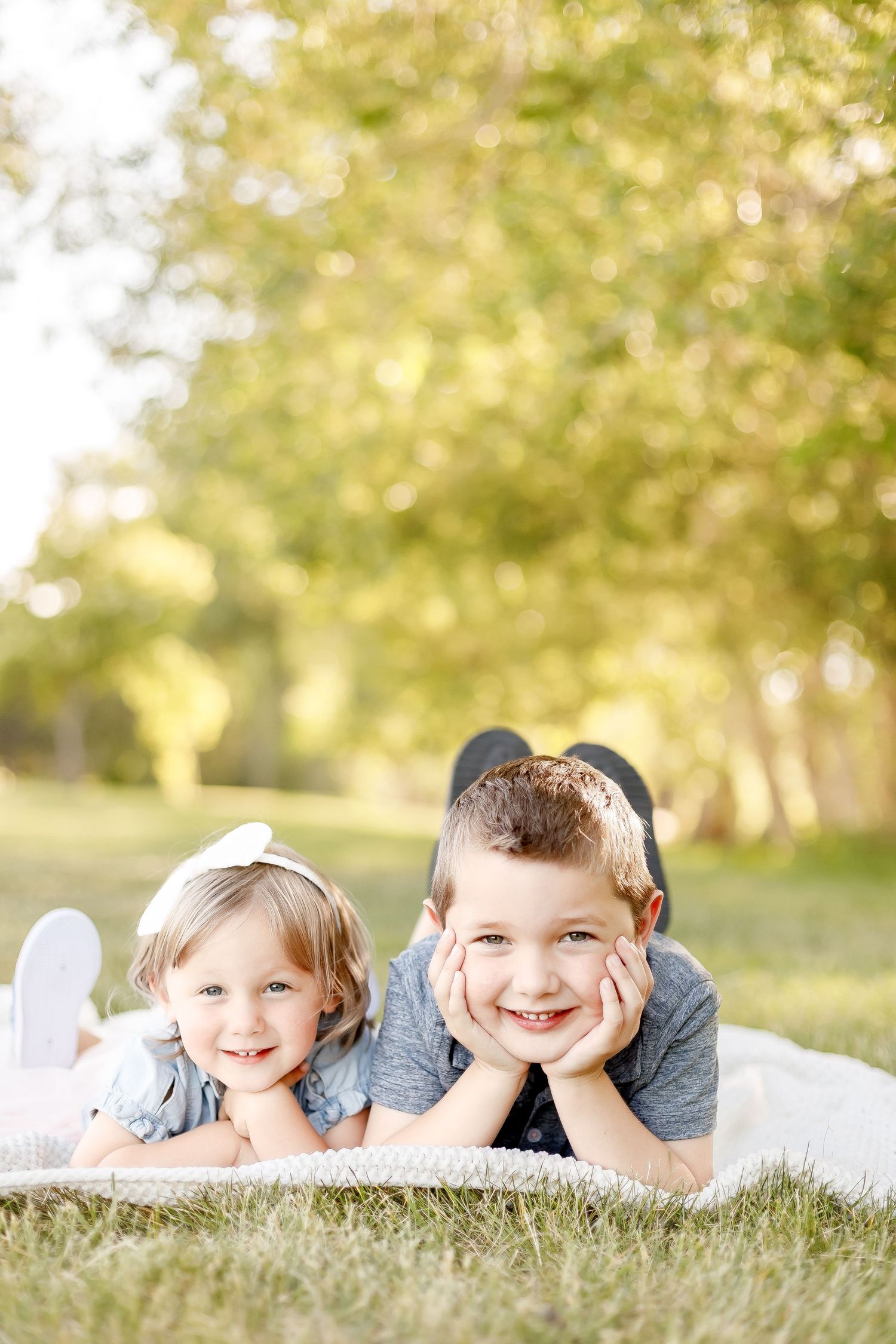 Two young siblings lie on a blanket in the grass smiling at the camera.