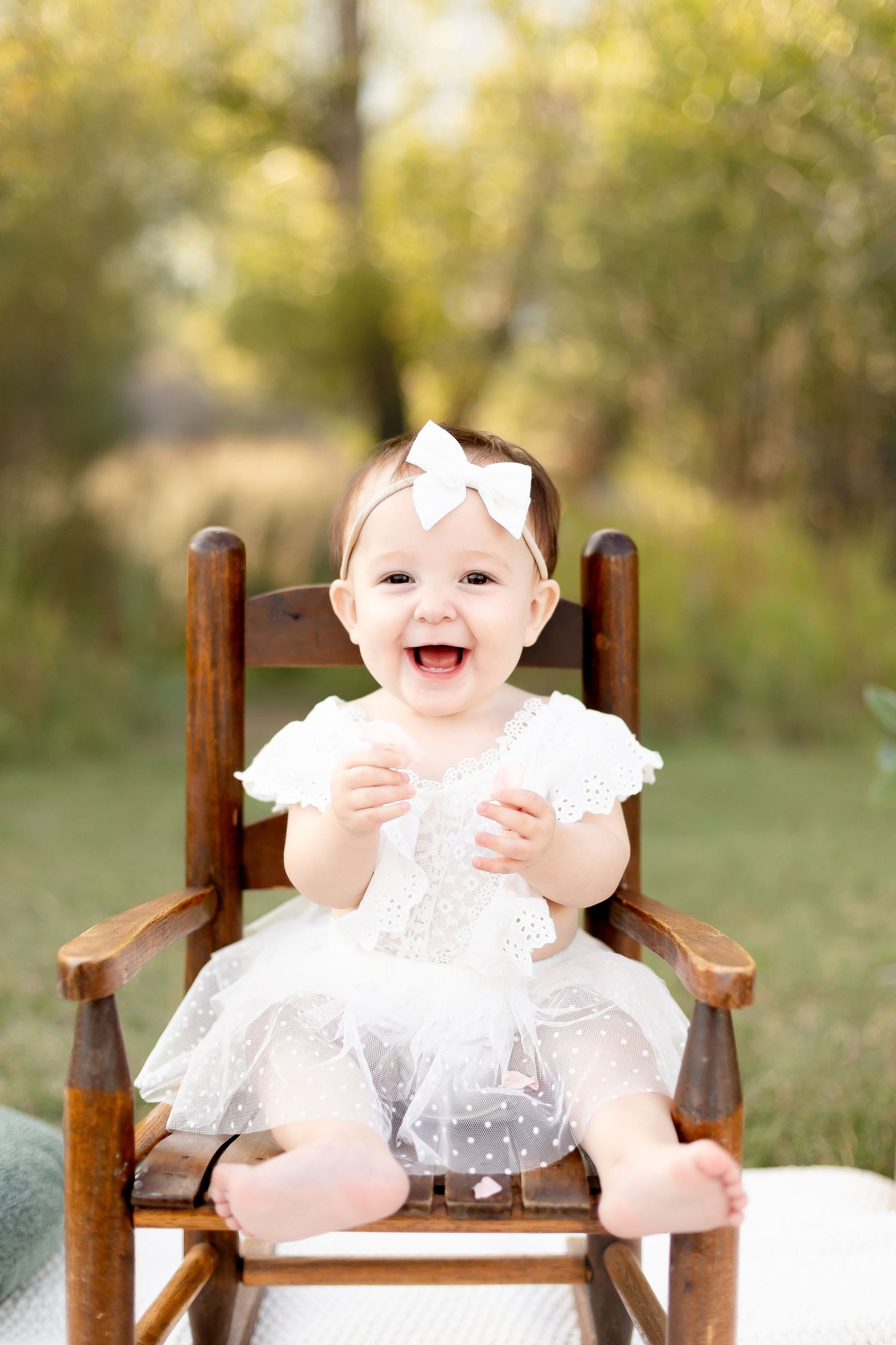 Laughing baby girl in white tulle dress with bow headband sitting in wooden rocking chair outdoors.
