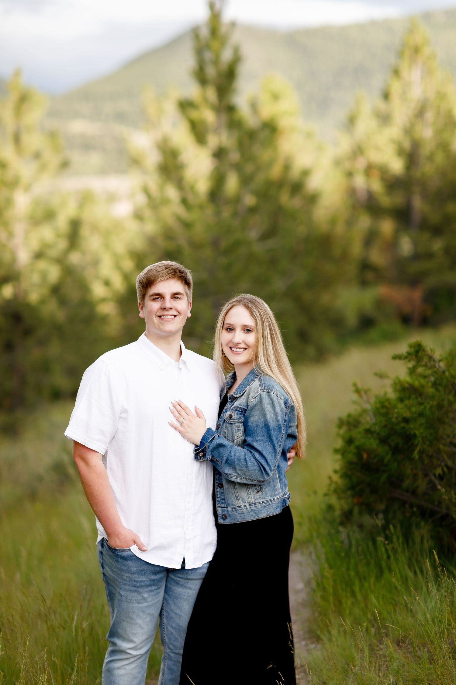 Smiling engaged couple posing together on a scenic mountain path surrounded by pine trees.