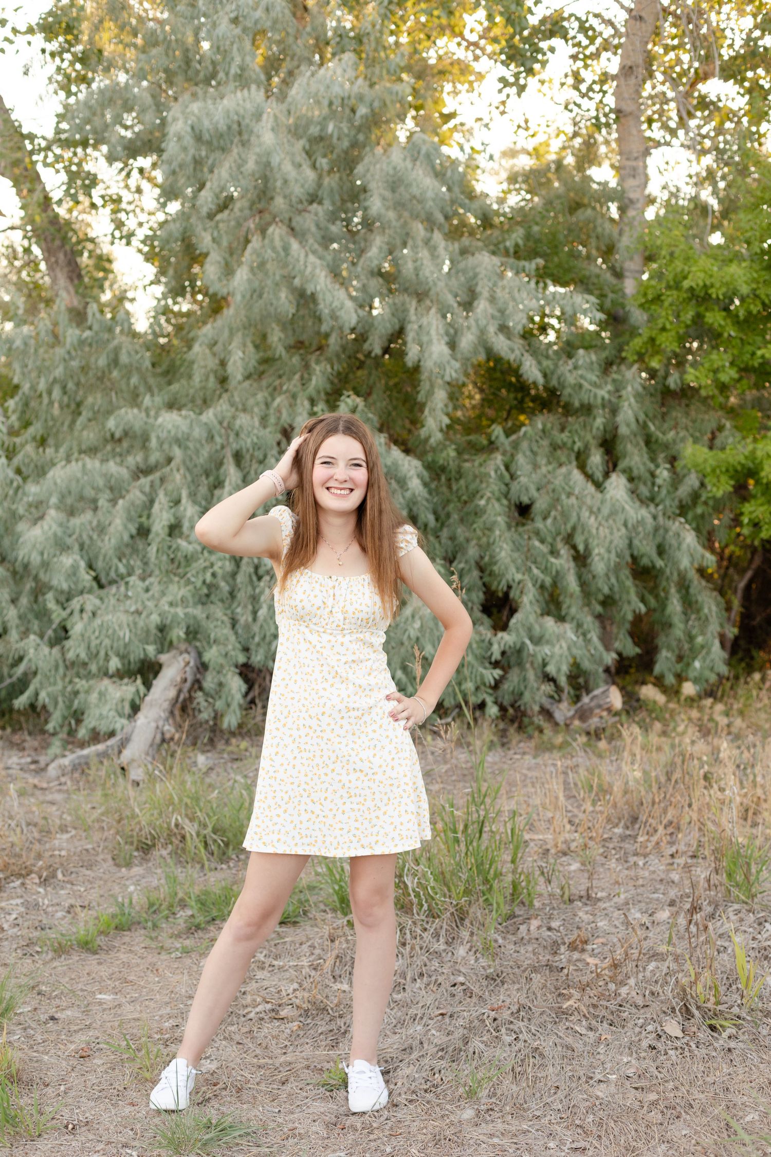 Smiling teenage girl in yellow floral sundress posing near green trees in natural setting.