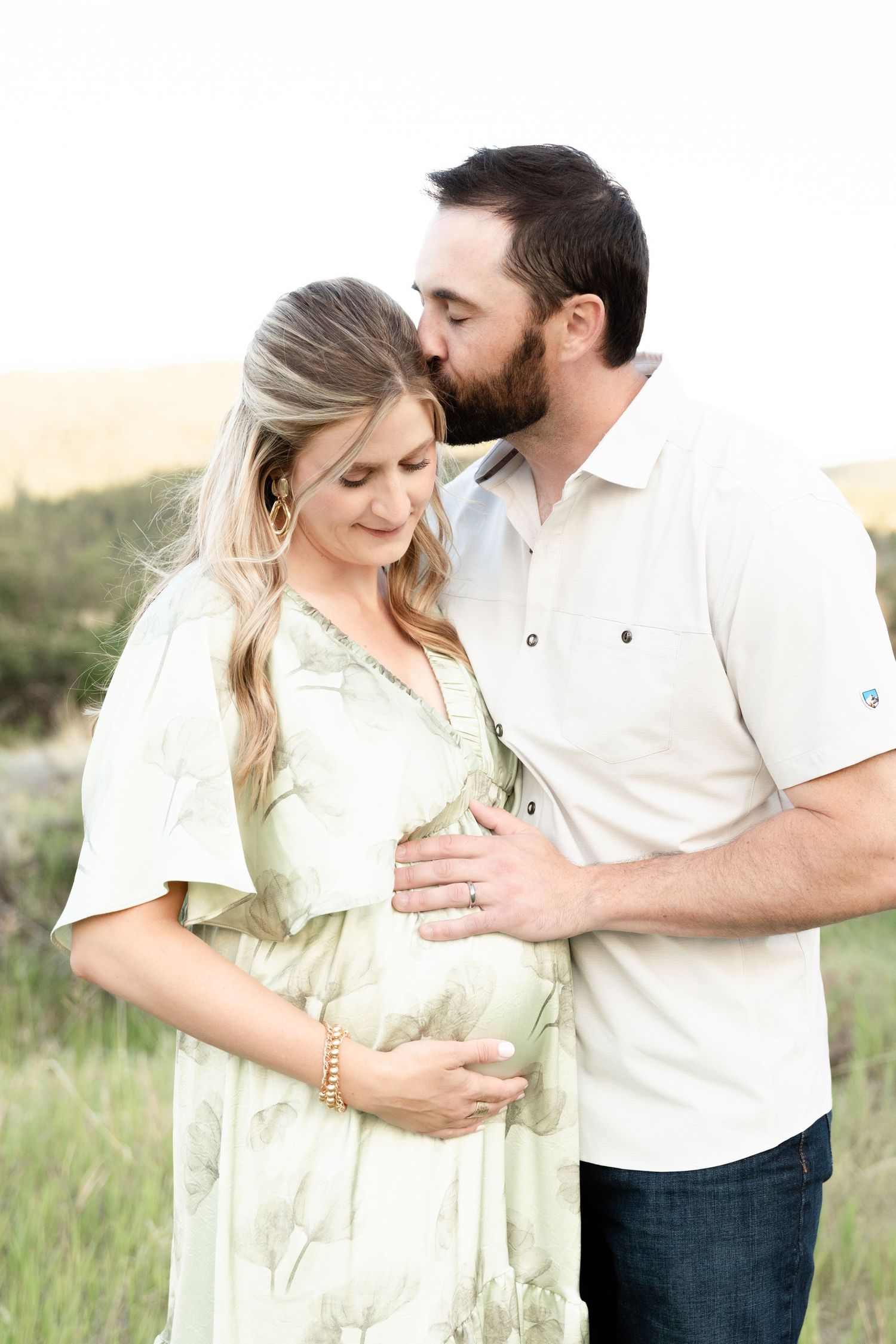 A bearded man tenderly kisses his pregnant wife's head while cradling her baby bump in a meadow.