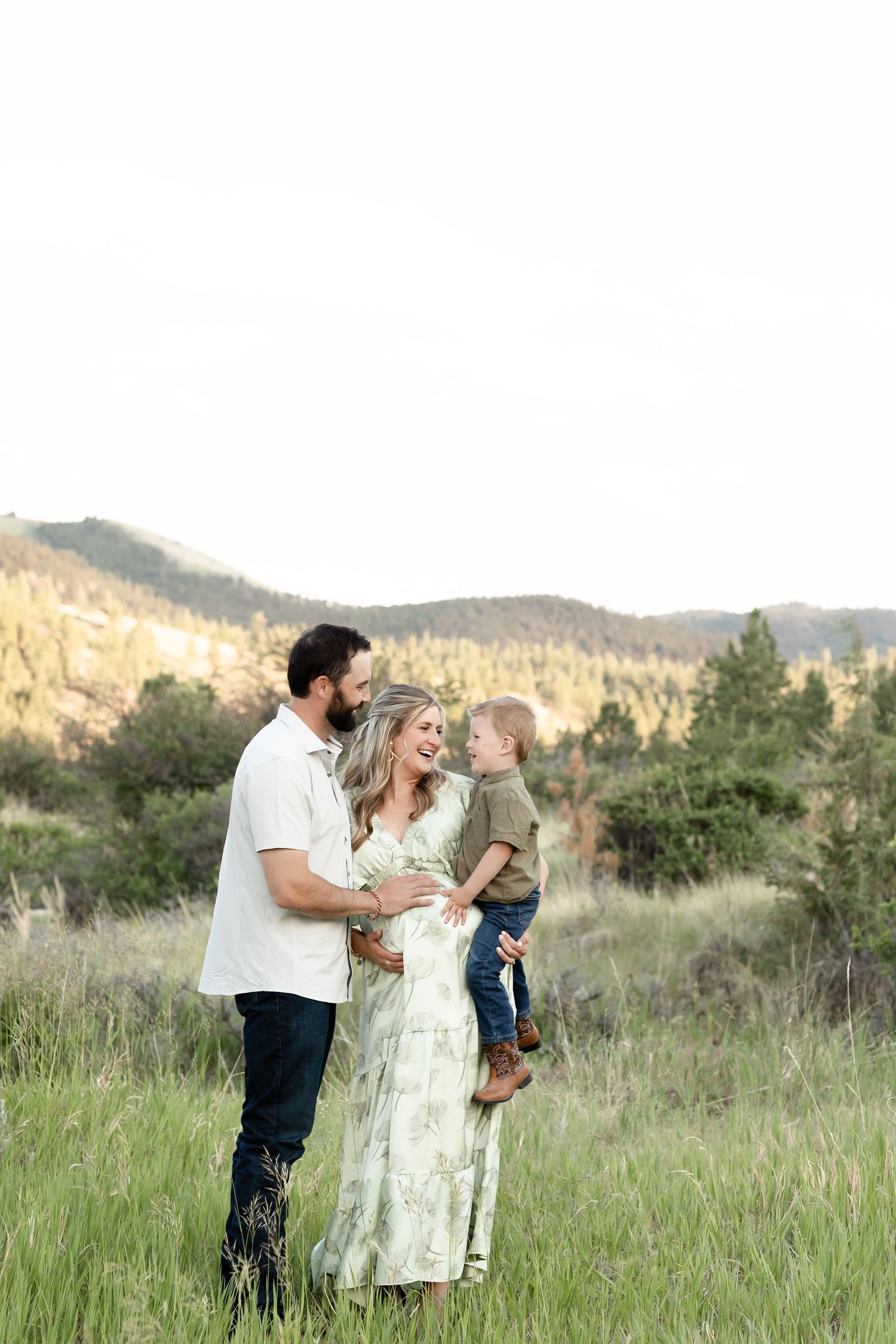 A happy expecting couple holds their young son in a lush green mountain meadow during a maternity shoot.