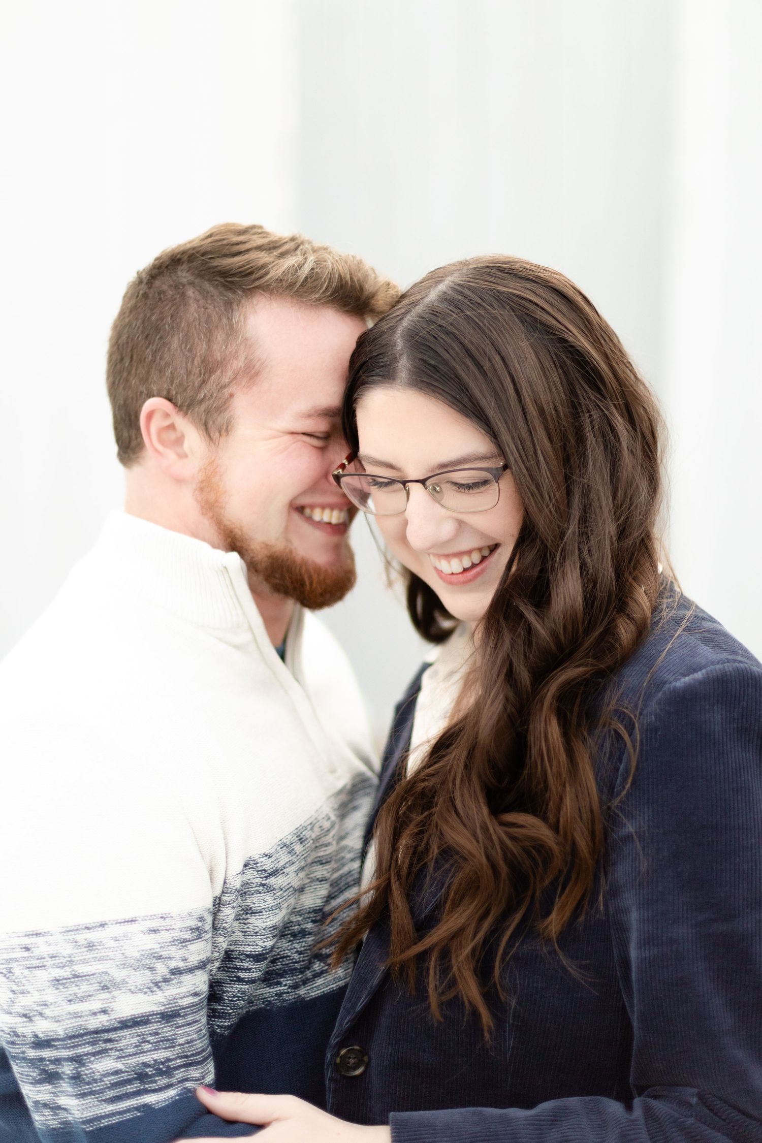 Happy couple laughing together indoors, man with red beard nuzzling brunette woman in glasses.
