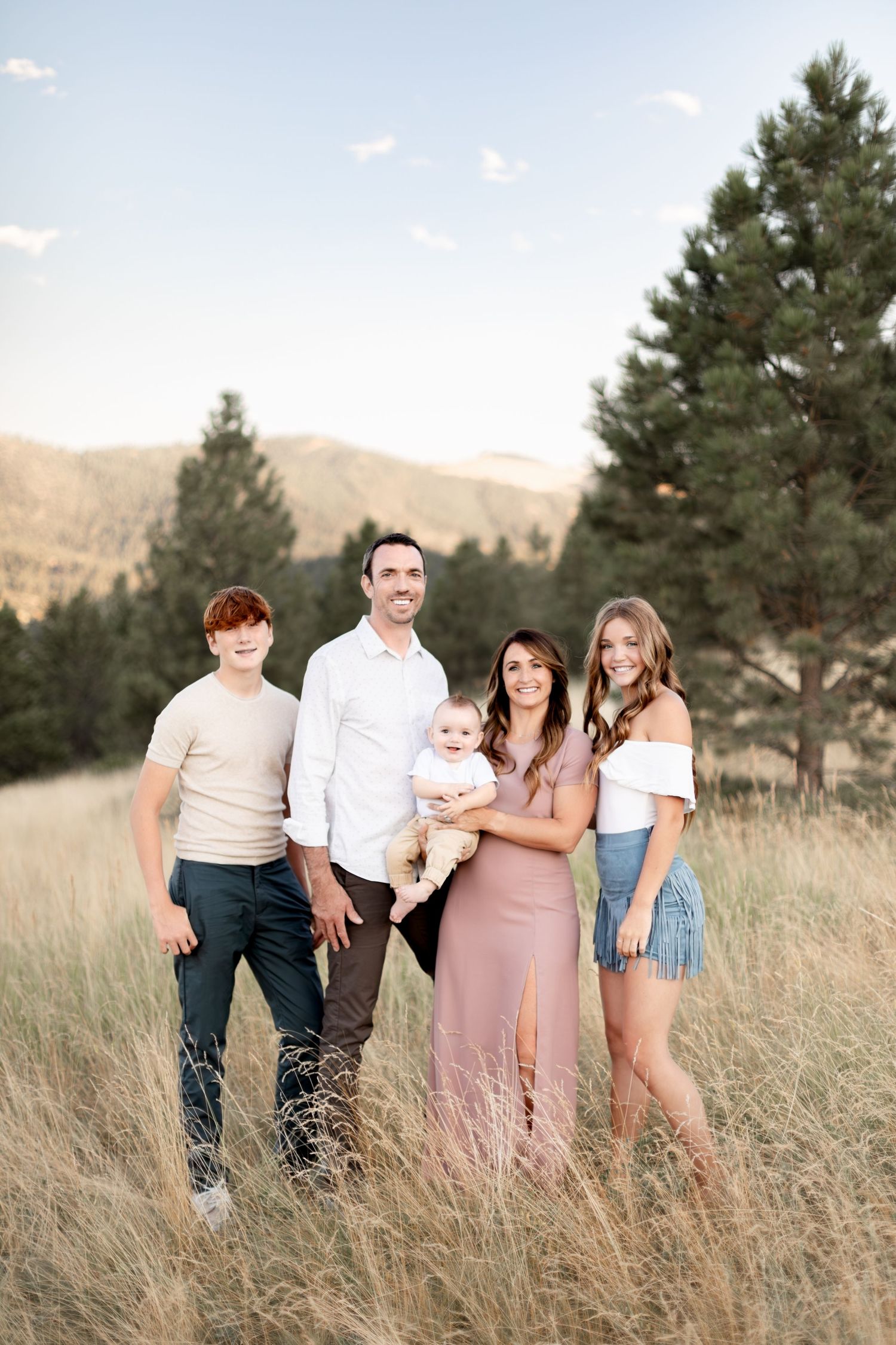 A family of five stands in a sunlit mountain meadow, mother holding a baby among tall grasses.