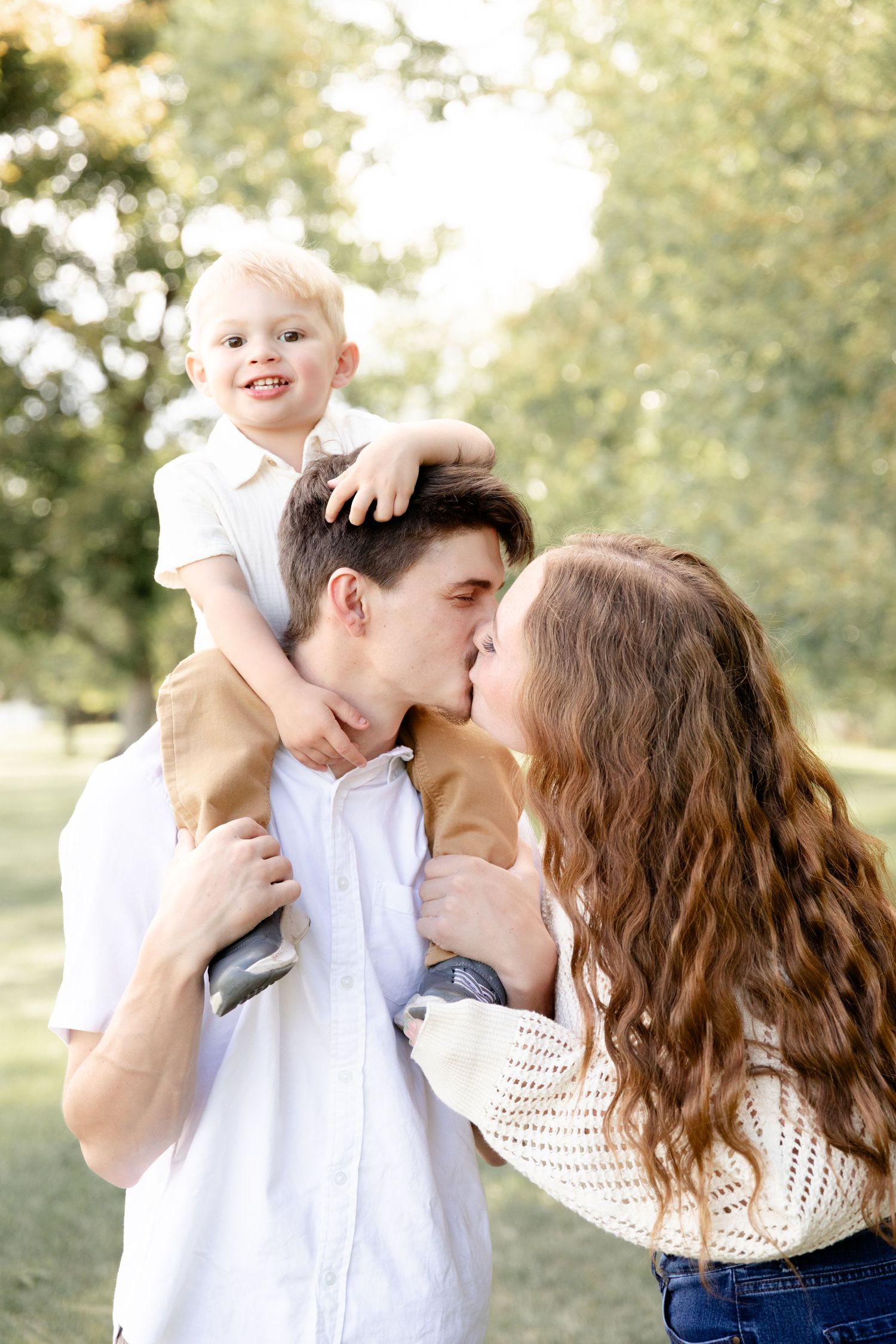 Young couple sharing a kiss while their smiling toddler sits on dads shoulders in a park.