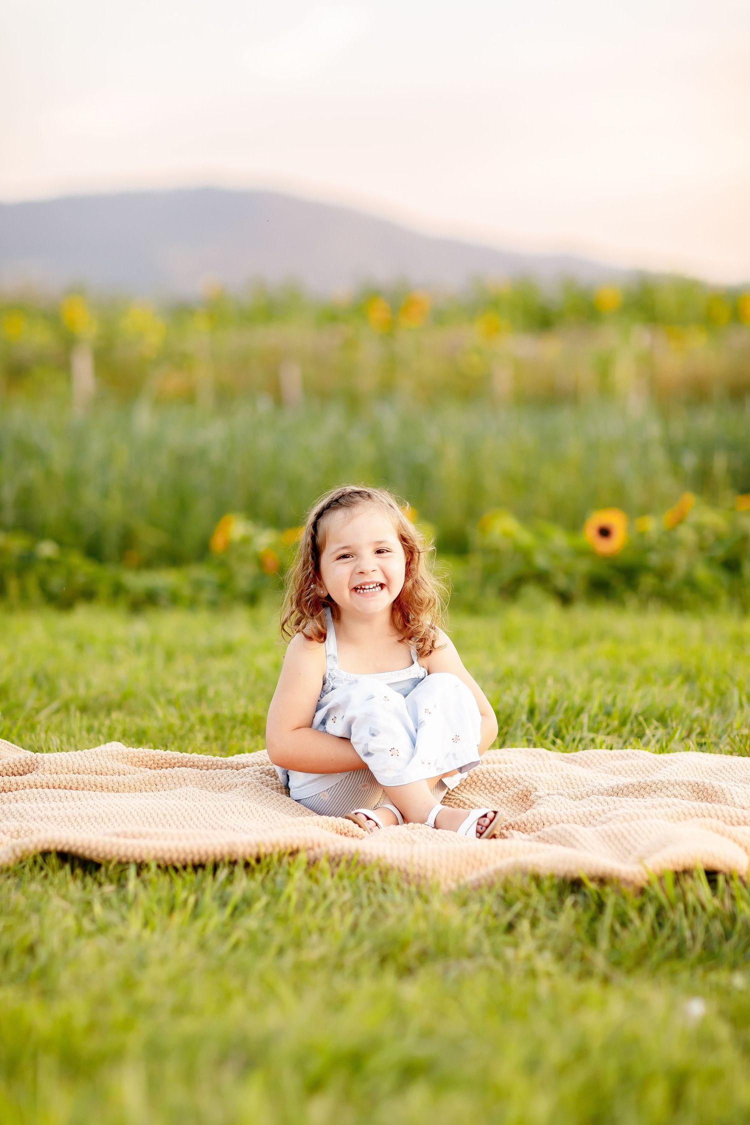 A happy young girl in a blue dress sits on a blanket in a sunflower field, smiling brightly on a warm day.