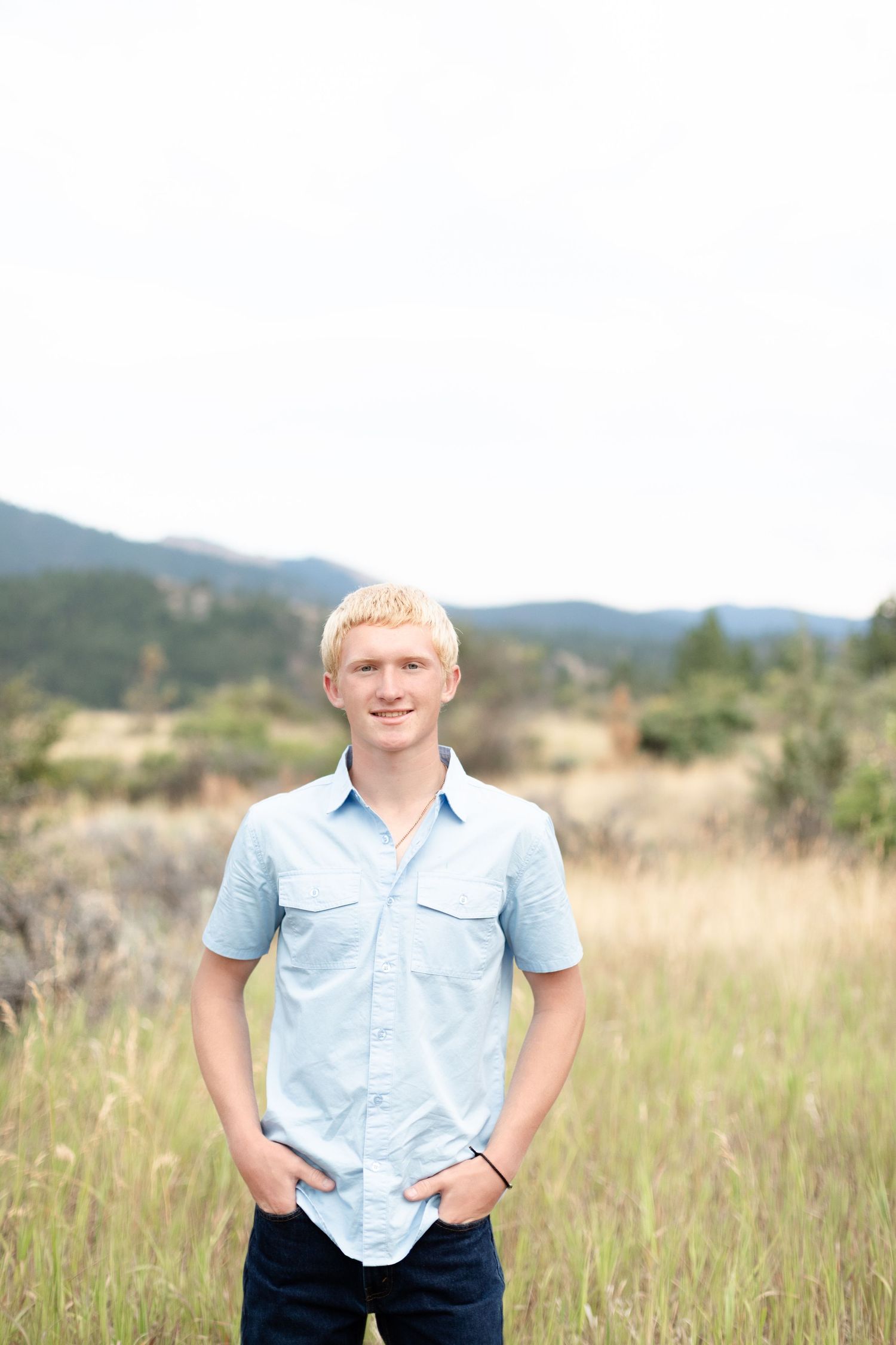 Teenage blonde boy in a light blue shirt smiling in a golden grass mountain field.