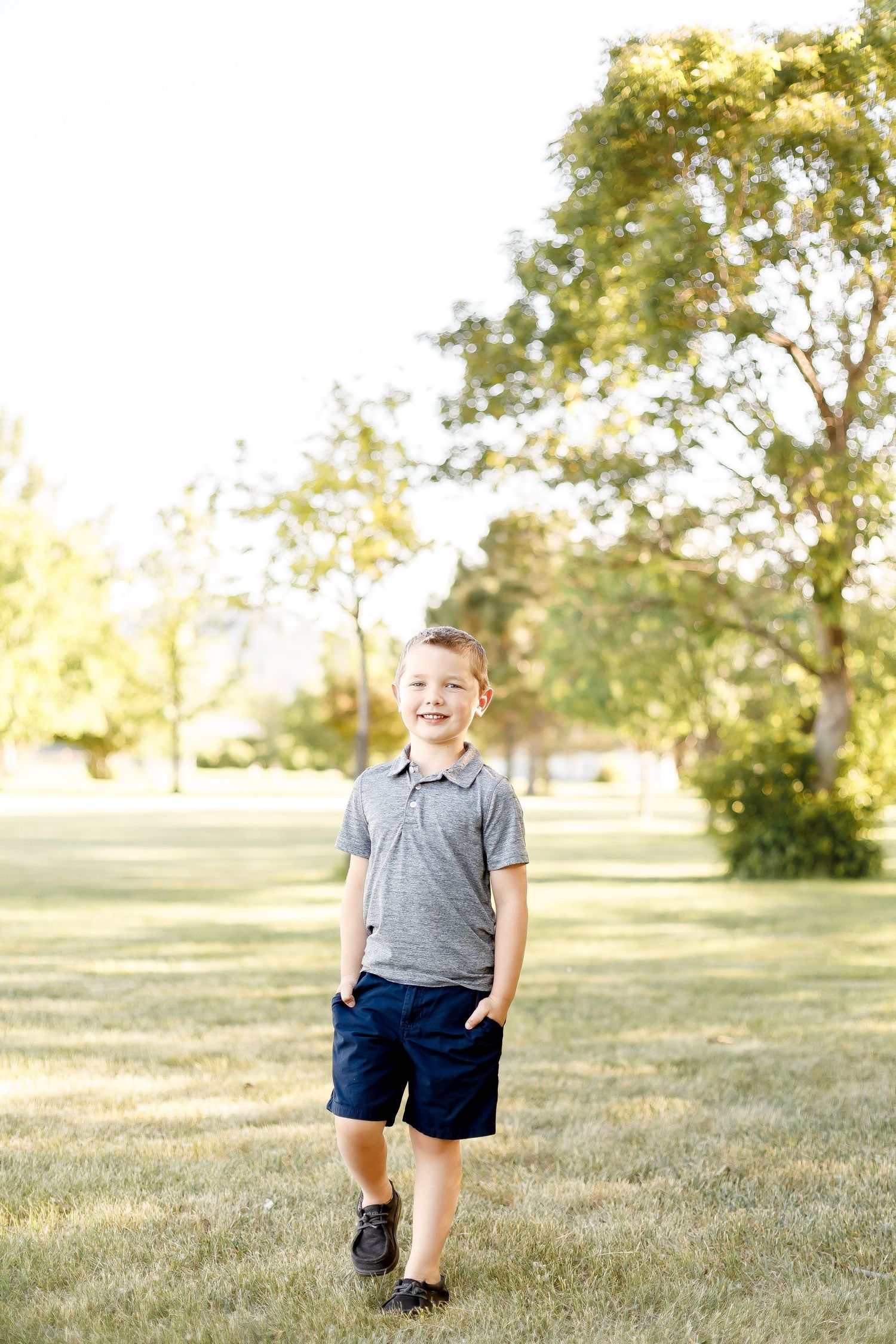 Young smiling boy with hands in pockets stands on green grass in a park.