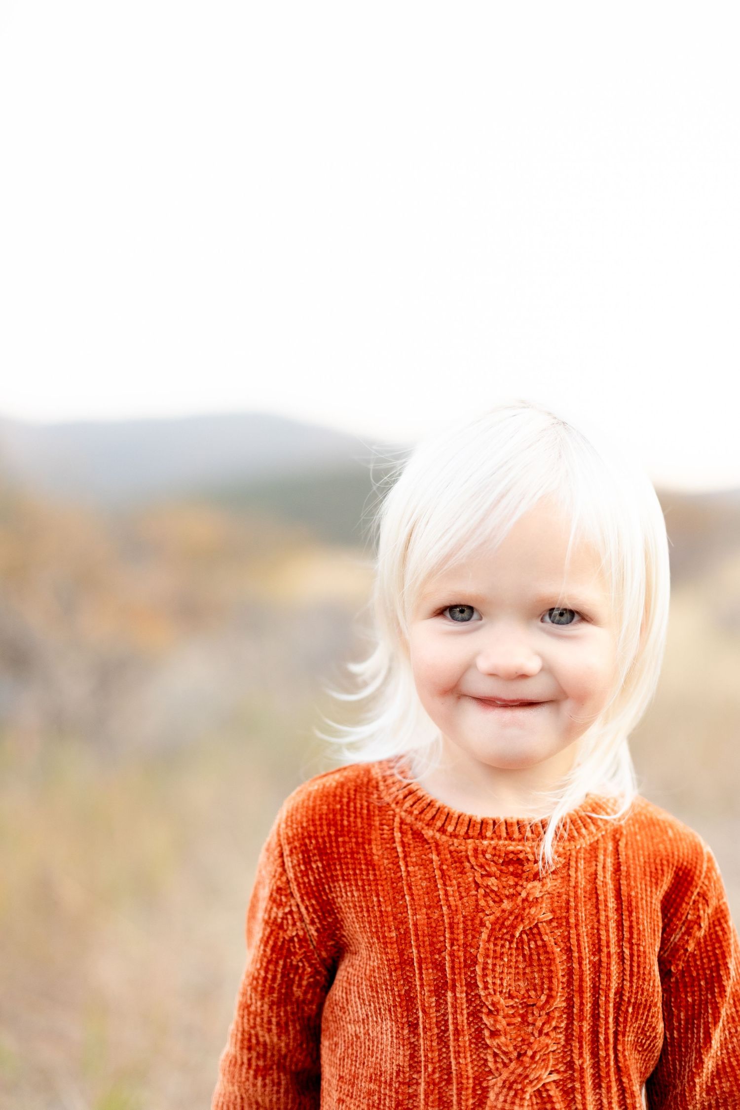 Close-up portrait of a cute blonde toddler girl in a rust orange cable-knit sweater smiling.