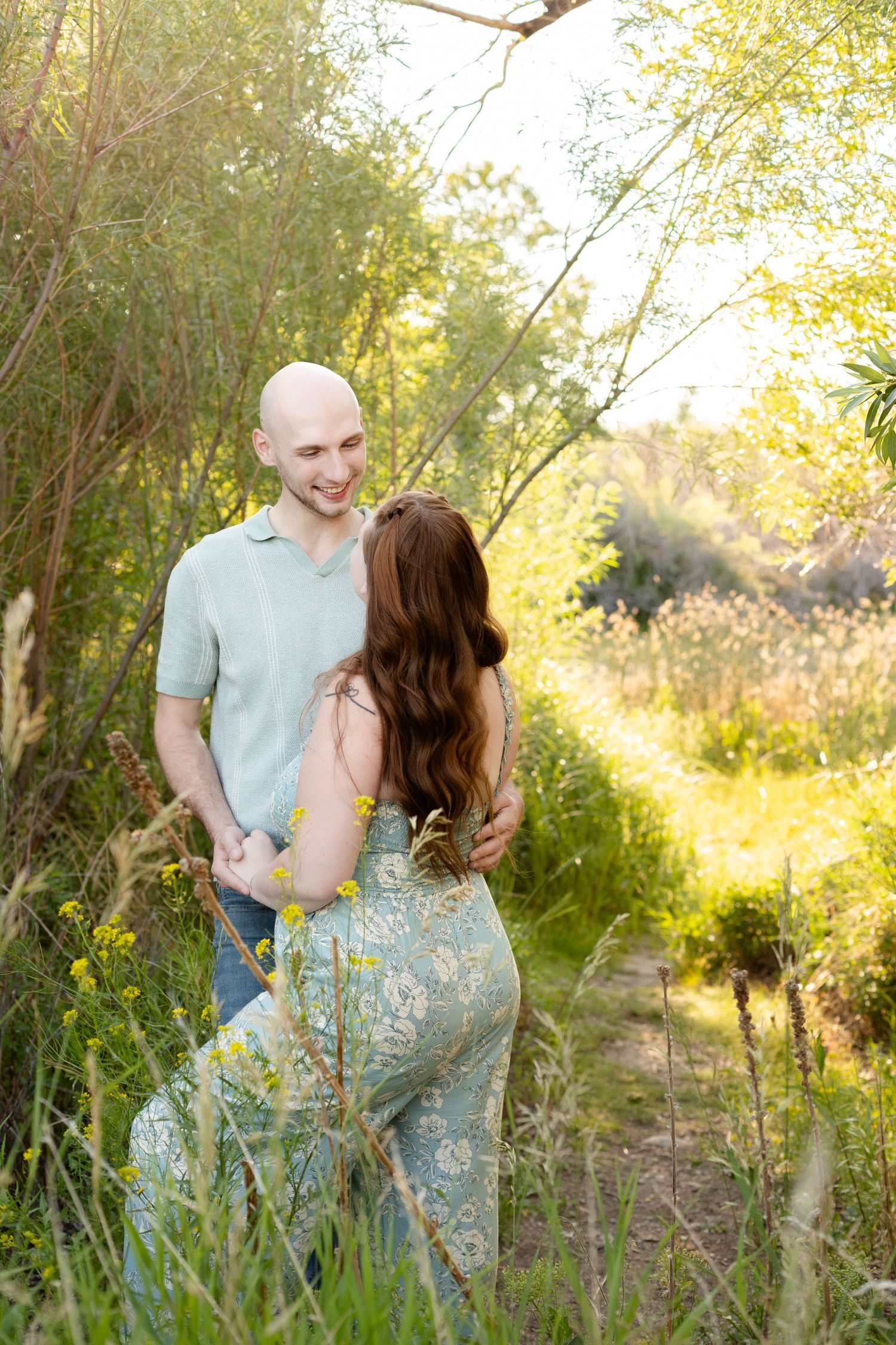 Bald man smiling at redhead woman holding hands in a lush green outdoor setting.