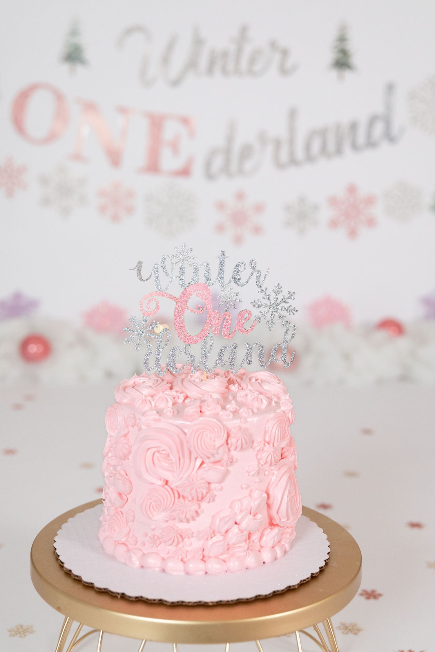 Pink rosette first birthday cake with Winter ONEderland topper on gold stand with snowflake decor.
