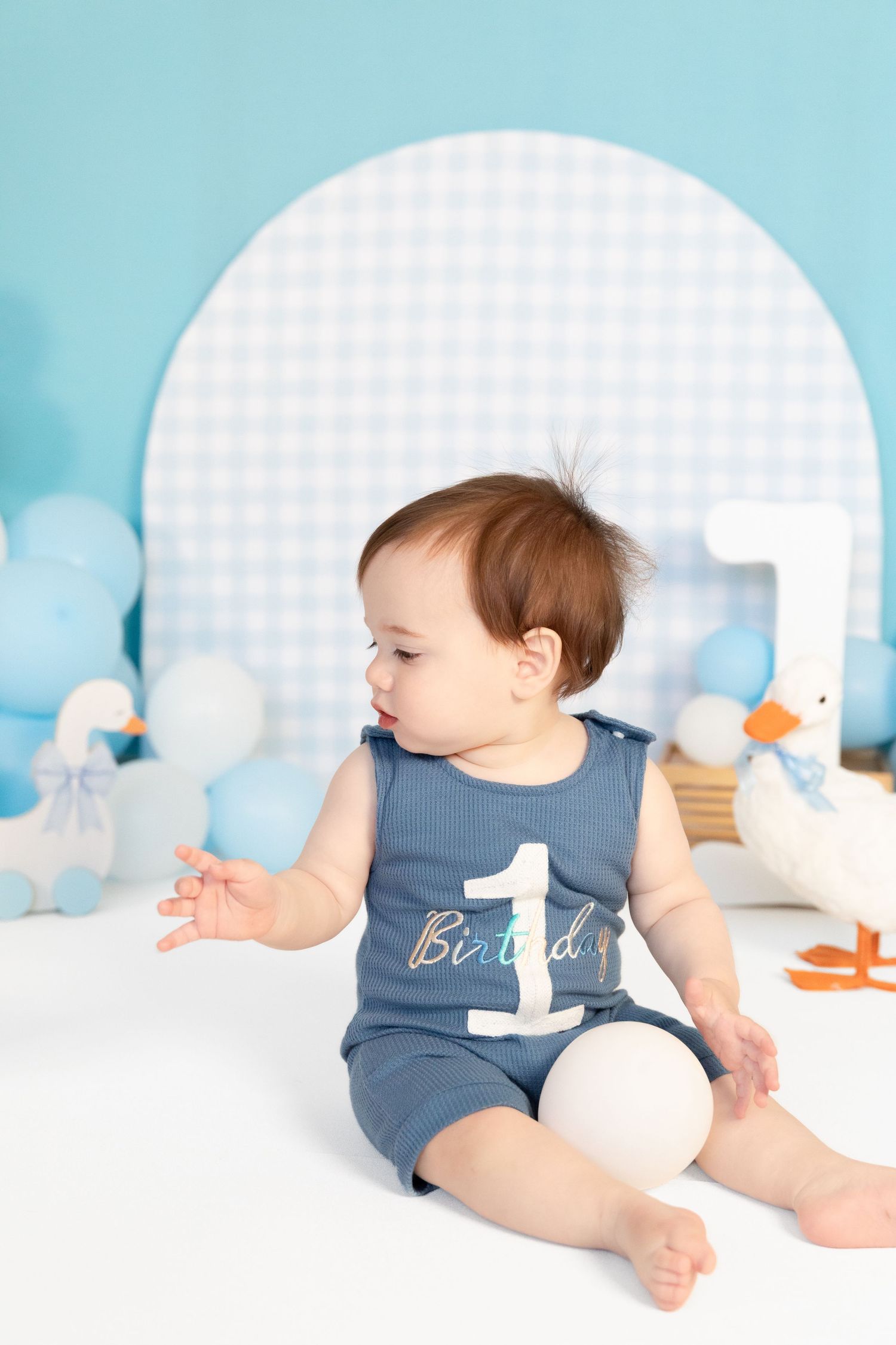 Baby boy in blue birthday romper sits among balloons and duck decorations at his first birthday photo shoot.