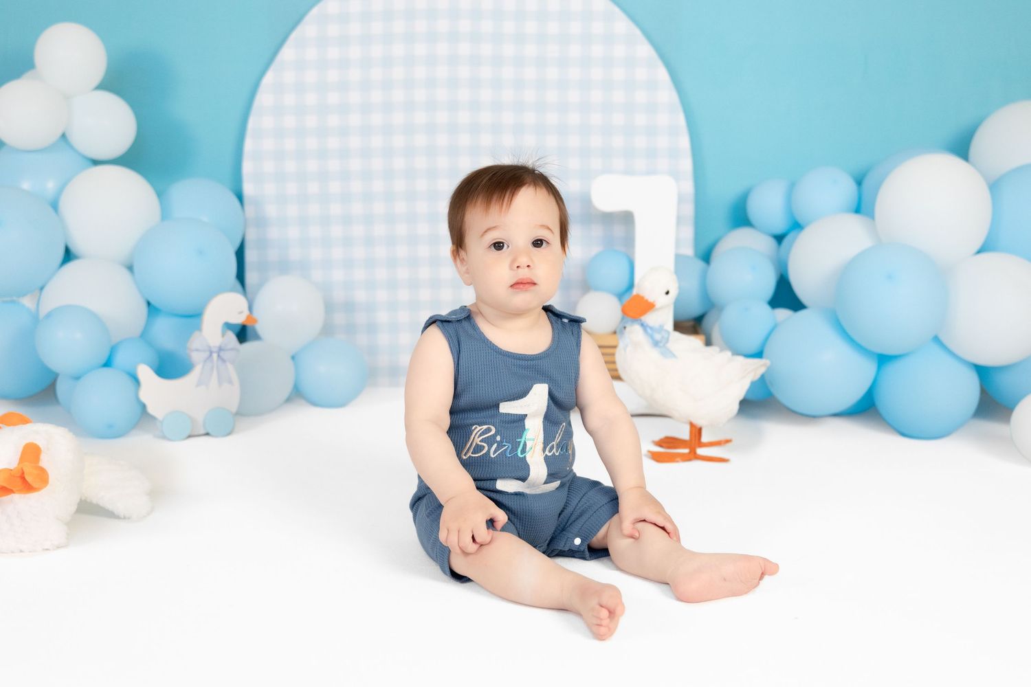 Adorable baby boy in a blue first birthday romper sitting among duck toys and blue balloons.
