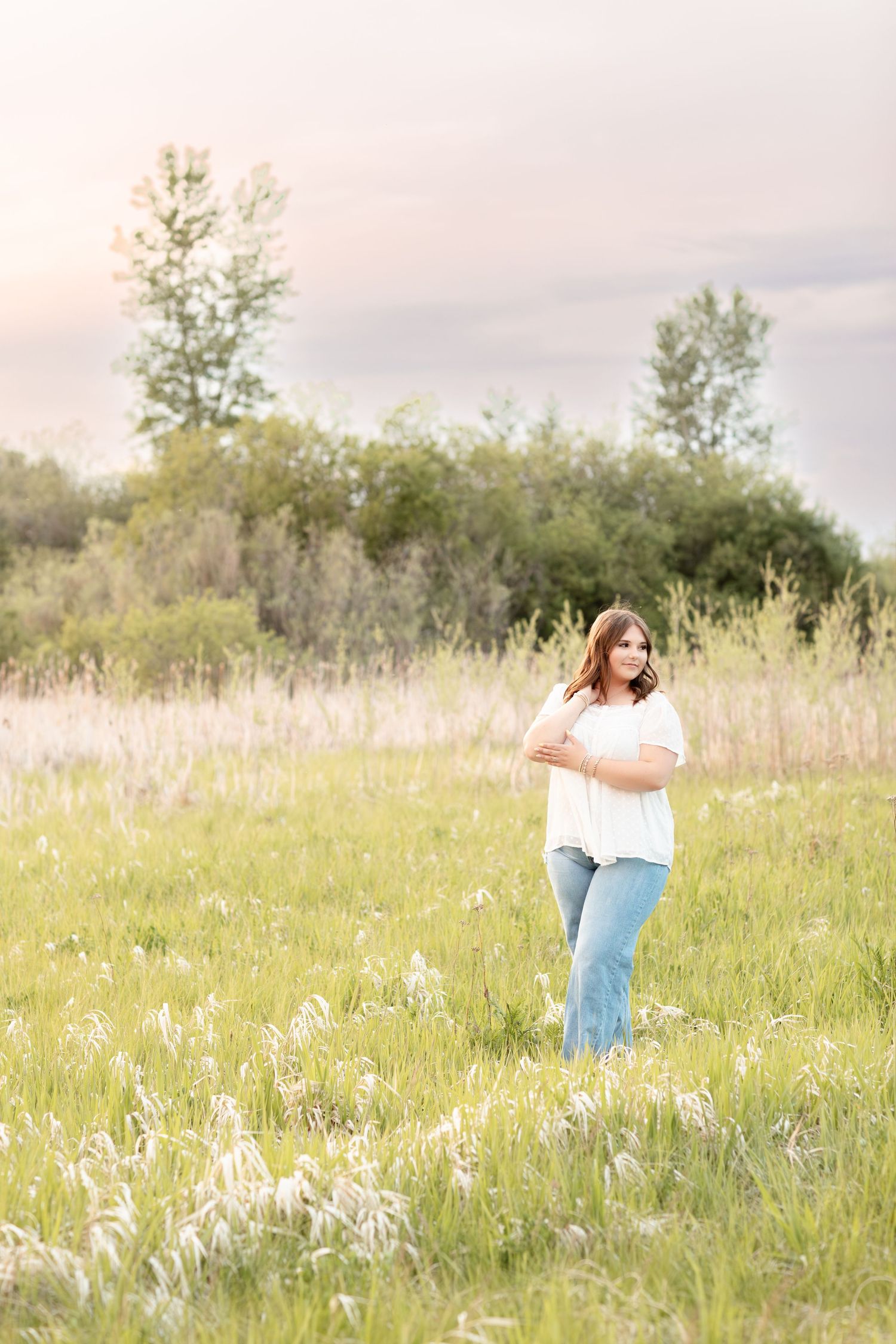 Young woman in white blouse and jeans standing in wildflower meadow at sunset.
