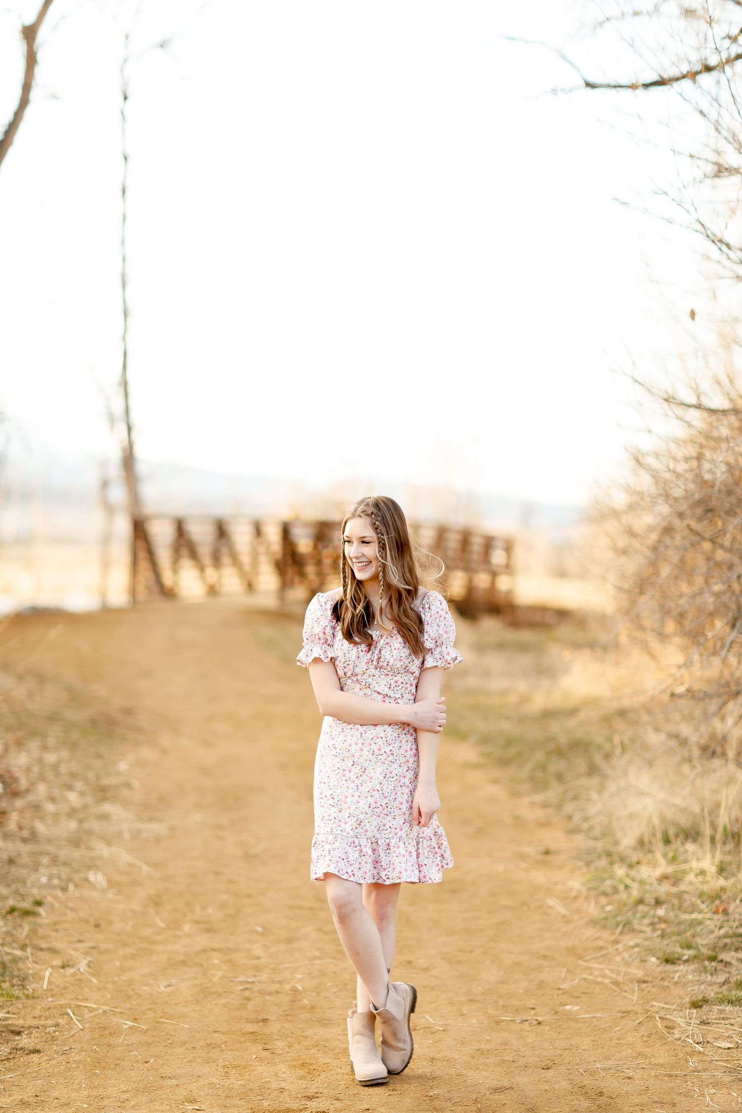 Young woman in floral dress and ankle boots walking on a dirt path with a wooden bridge in the background.