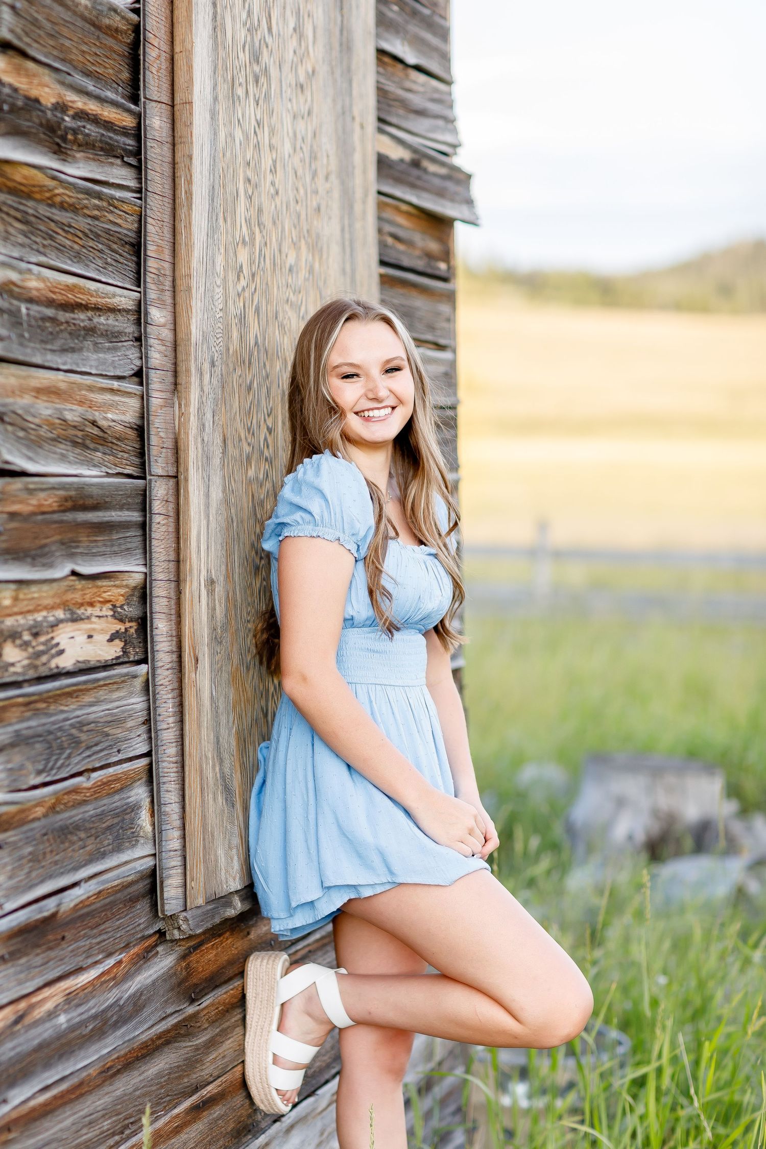 Smiling teenage girl in a light blue dress leaning against a rustic wooden barn wall.