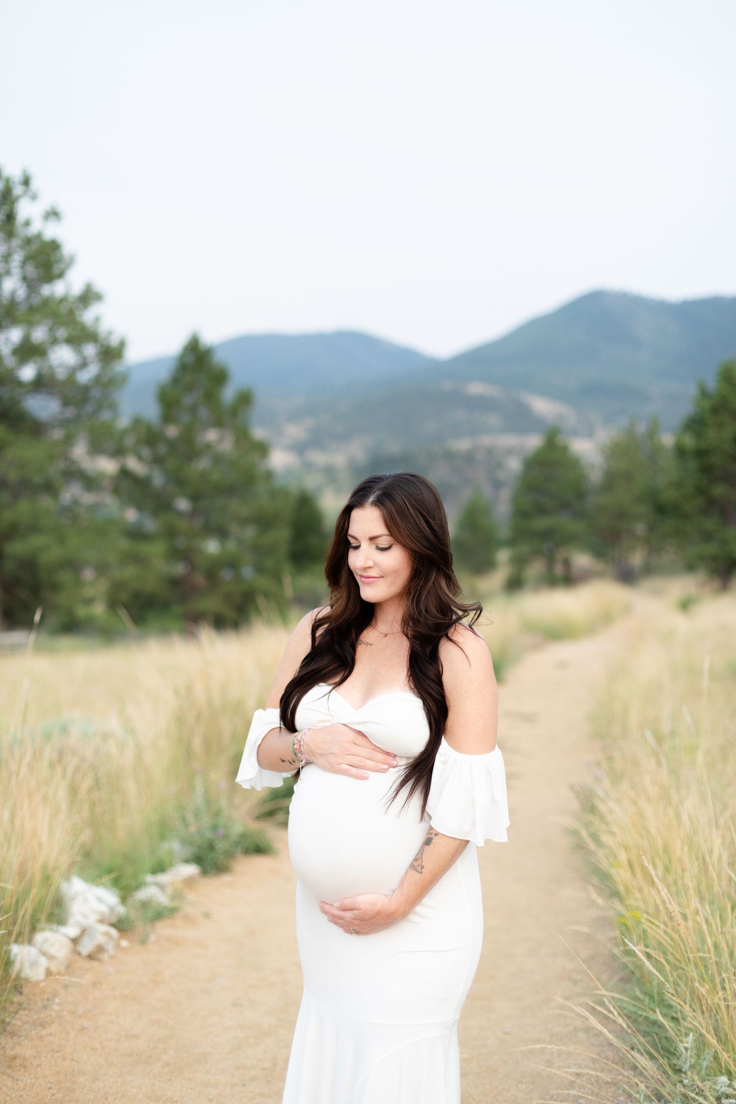 Pregnant woman in white off-shoulder dress gazing down at baby bump on trail.