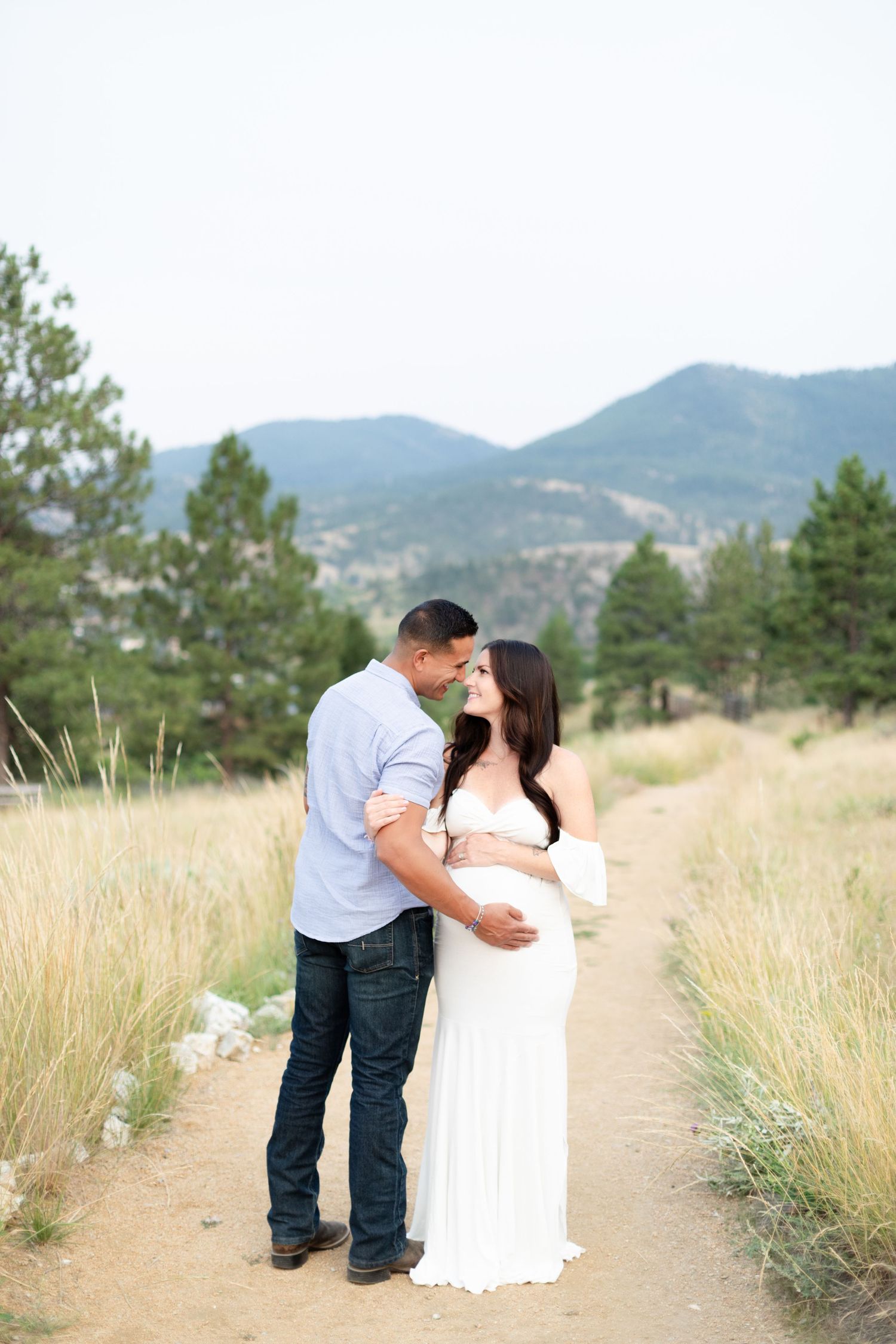 Couple sharing a romantic moment on a dirt path surrounded by tall grass and mountain scenery.