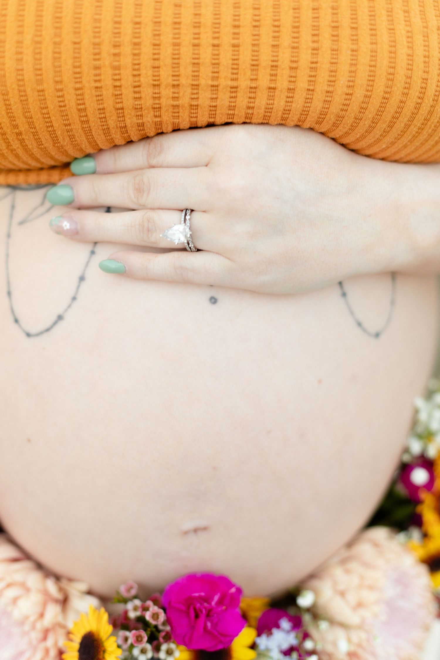 Close-up of pregnant belly with tattoos, floral bouquet, and ring on hand wearing orange top.