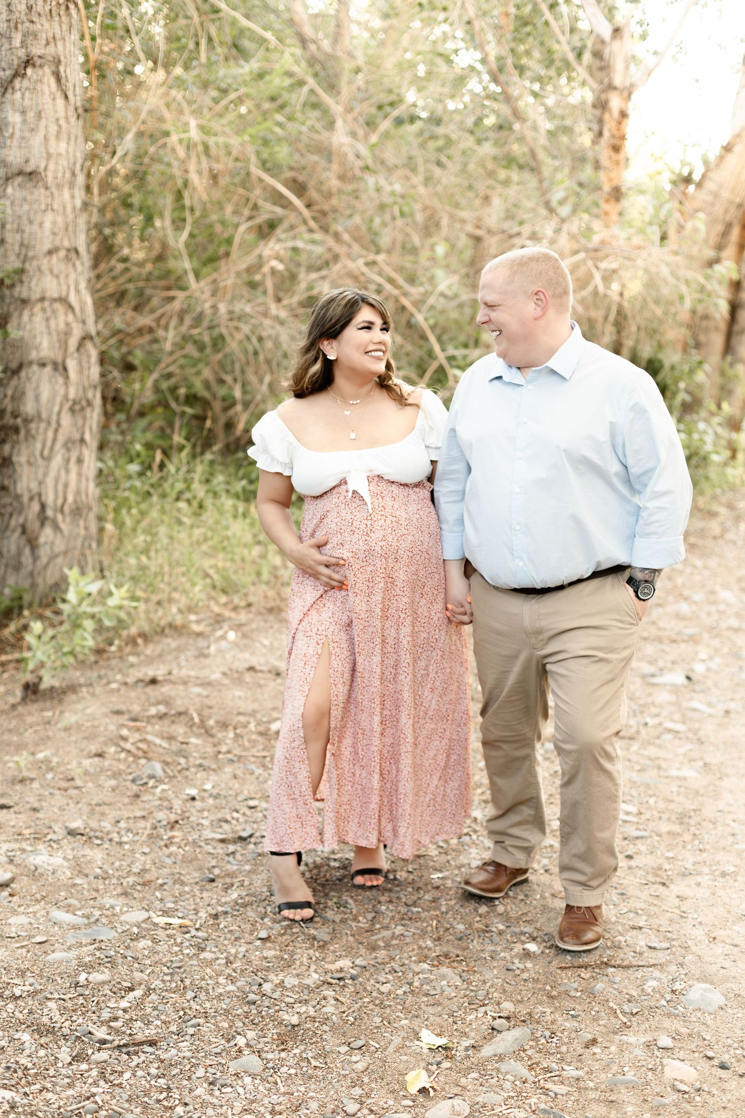 Smiling pregnant woman in floral dress walks with partner on gravel path during outdoor couples maternity photo session.