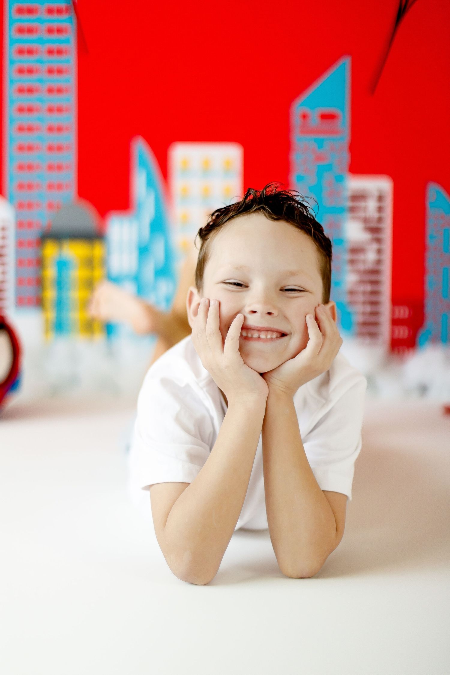 A smiling boy lies on the floor in front of a colorful Spiderman-themed cityscape backdrop.