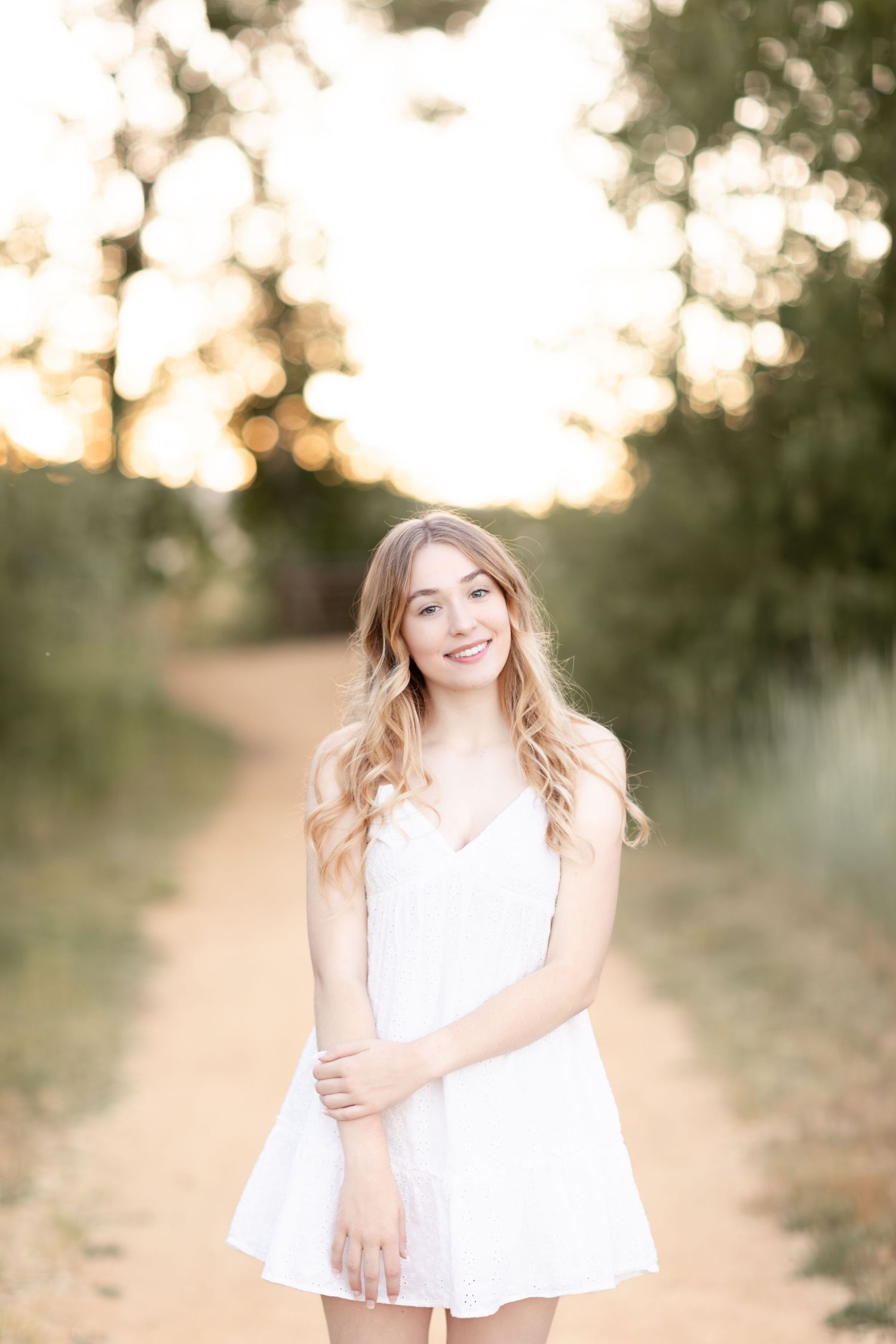 Smiling young woman in white dress standing on sunlit dirt path with bokeh trees in warm golden hour light.