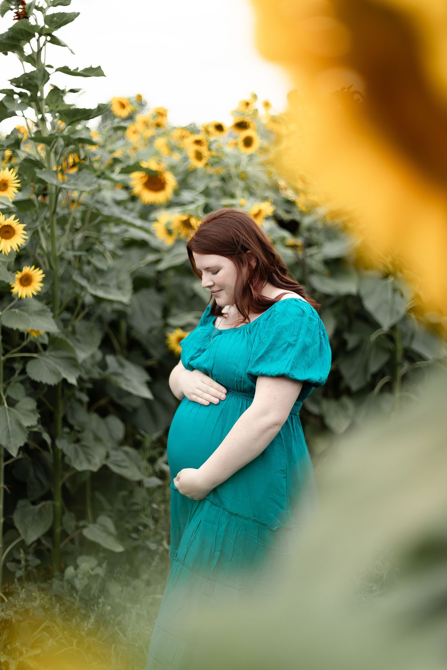 Pregnant woman in teal dress gazes down at baby bump surrounded by blooming sunflowers outdoors.