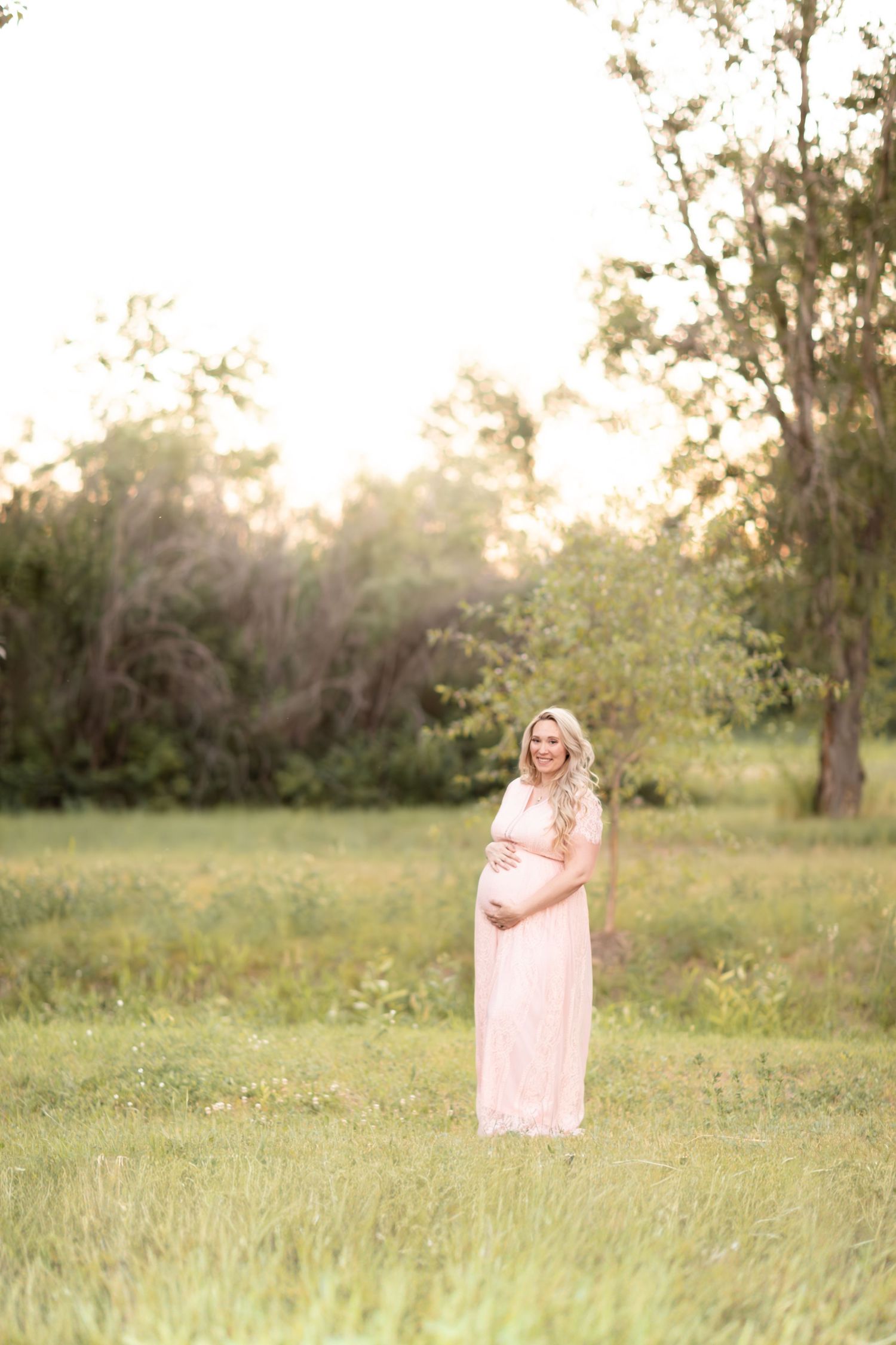 Smiling pregnant woman in blush pink lace maternity gown standing in green field at golden hour.