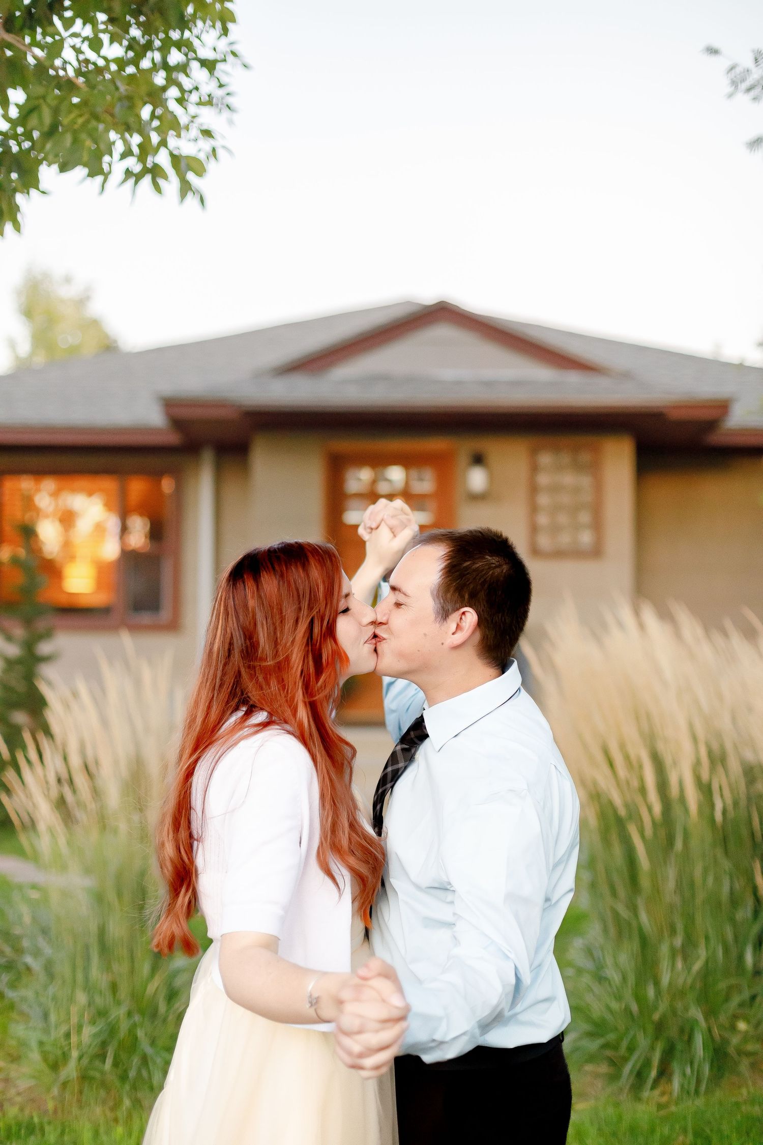 Engaged couple sharing a kiss while dancing outdoors in front of a house at dusk.