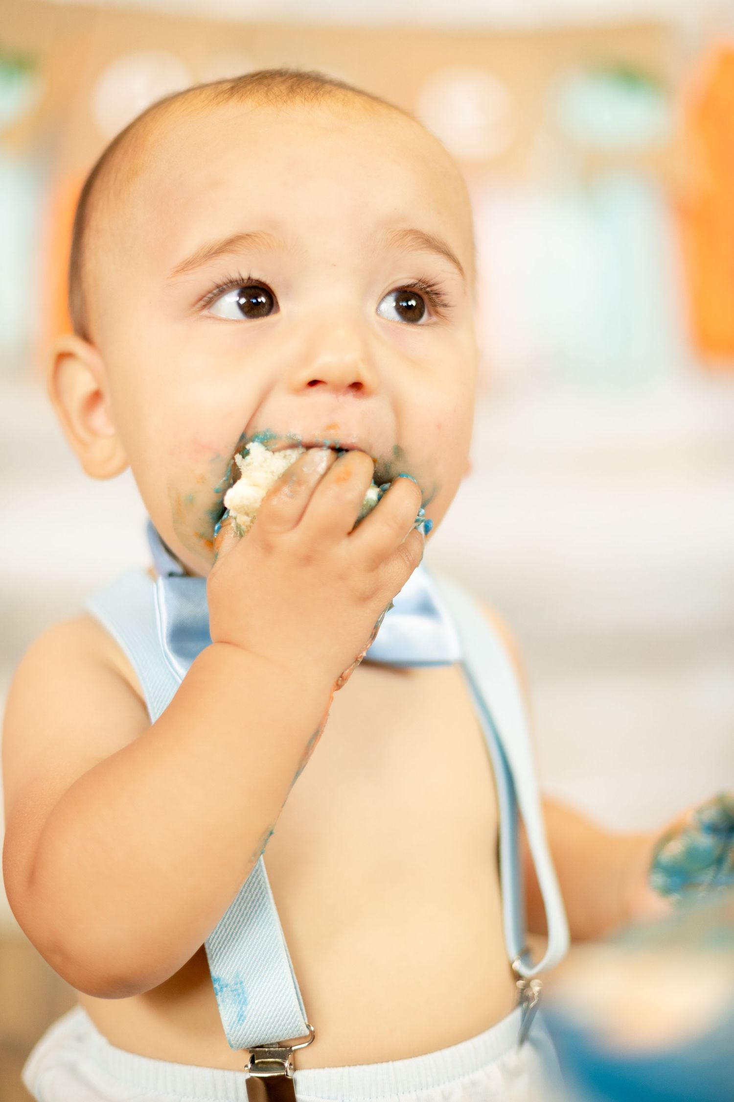 Messy baby boy in blue bow tie suspenders eating birthday cake with hand at first birthday cake smash.