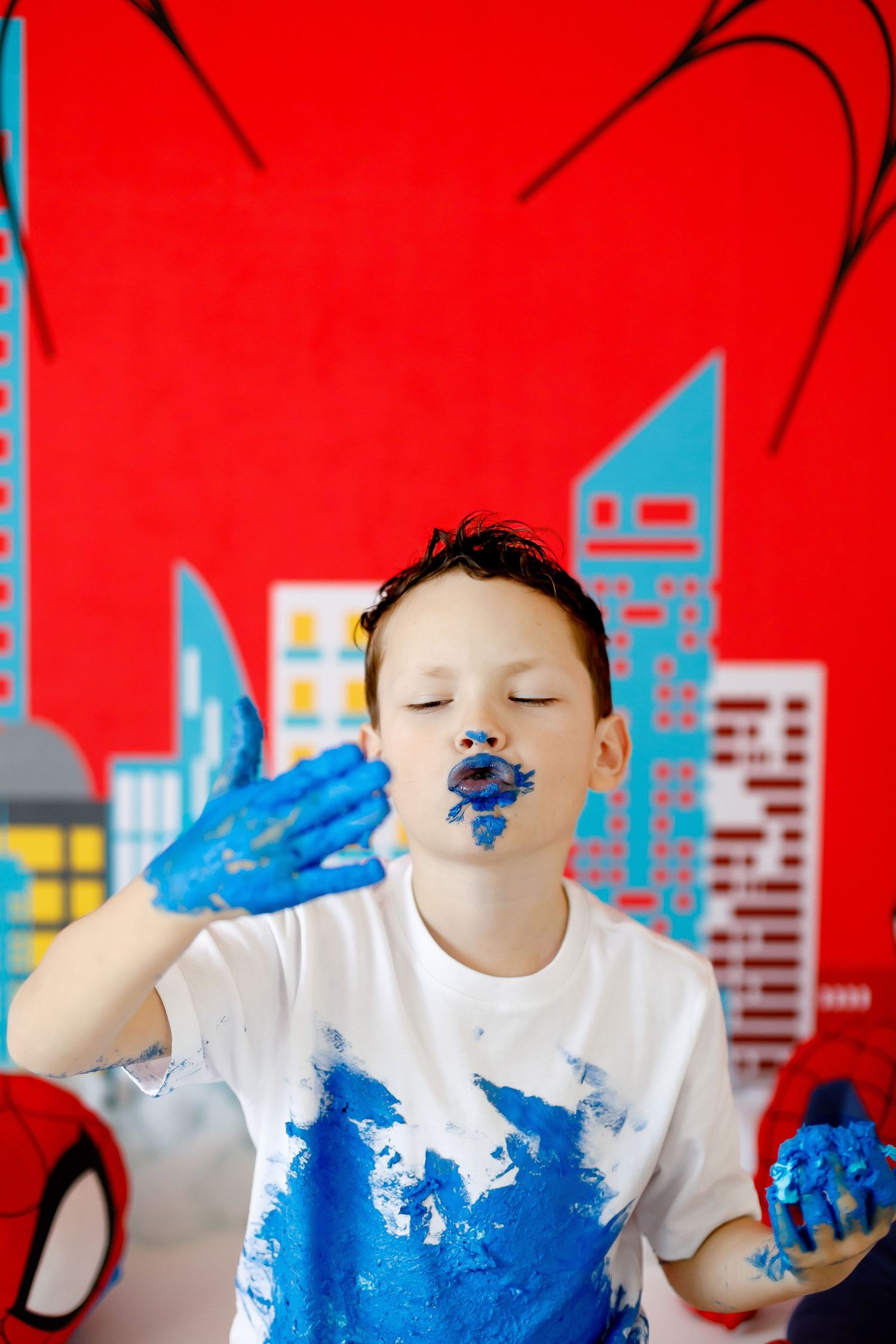 A boy with blue cake frosting on his face blows a kiss at a Spiderman birthday party.