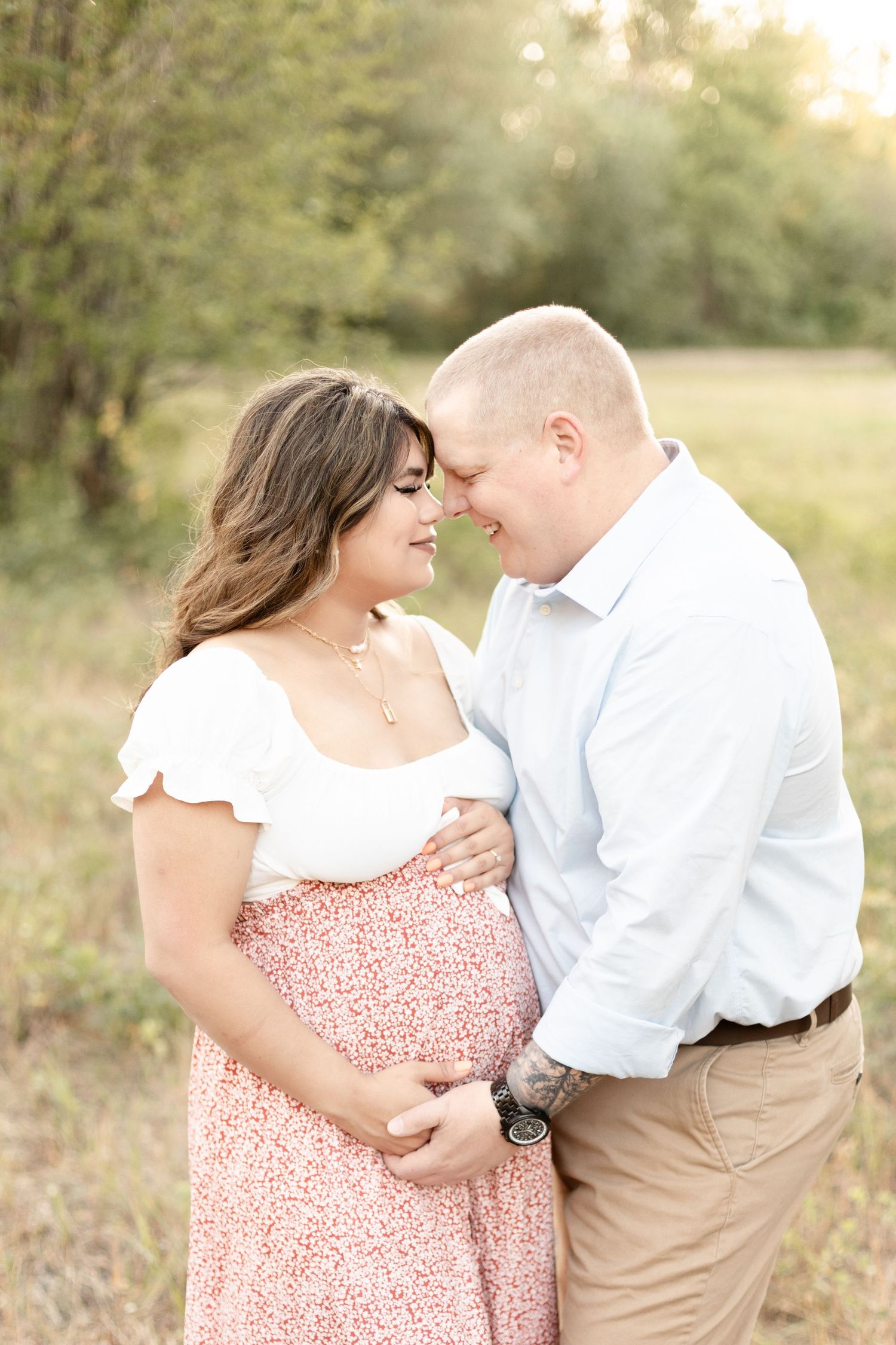 Couple sharing a tender forehead touch outdoors, woman in floral maternity dress holding baby bump in golden light.