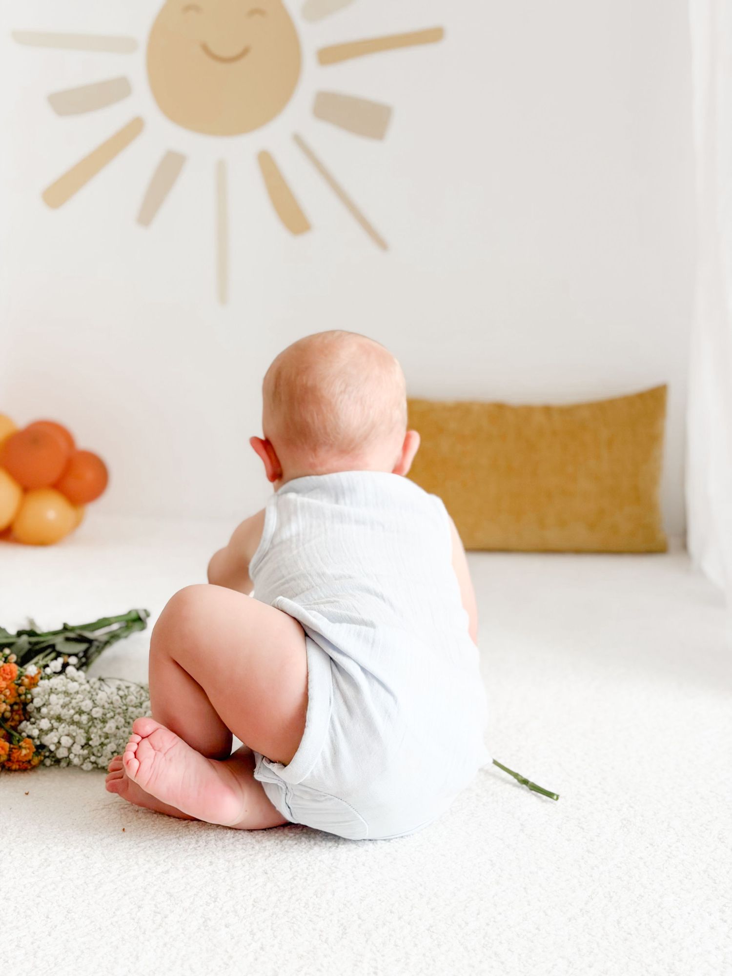 Baby in white romper sitting facing away near flowers in bright sun-themed studio setting.