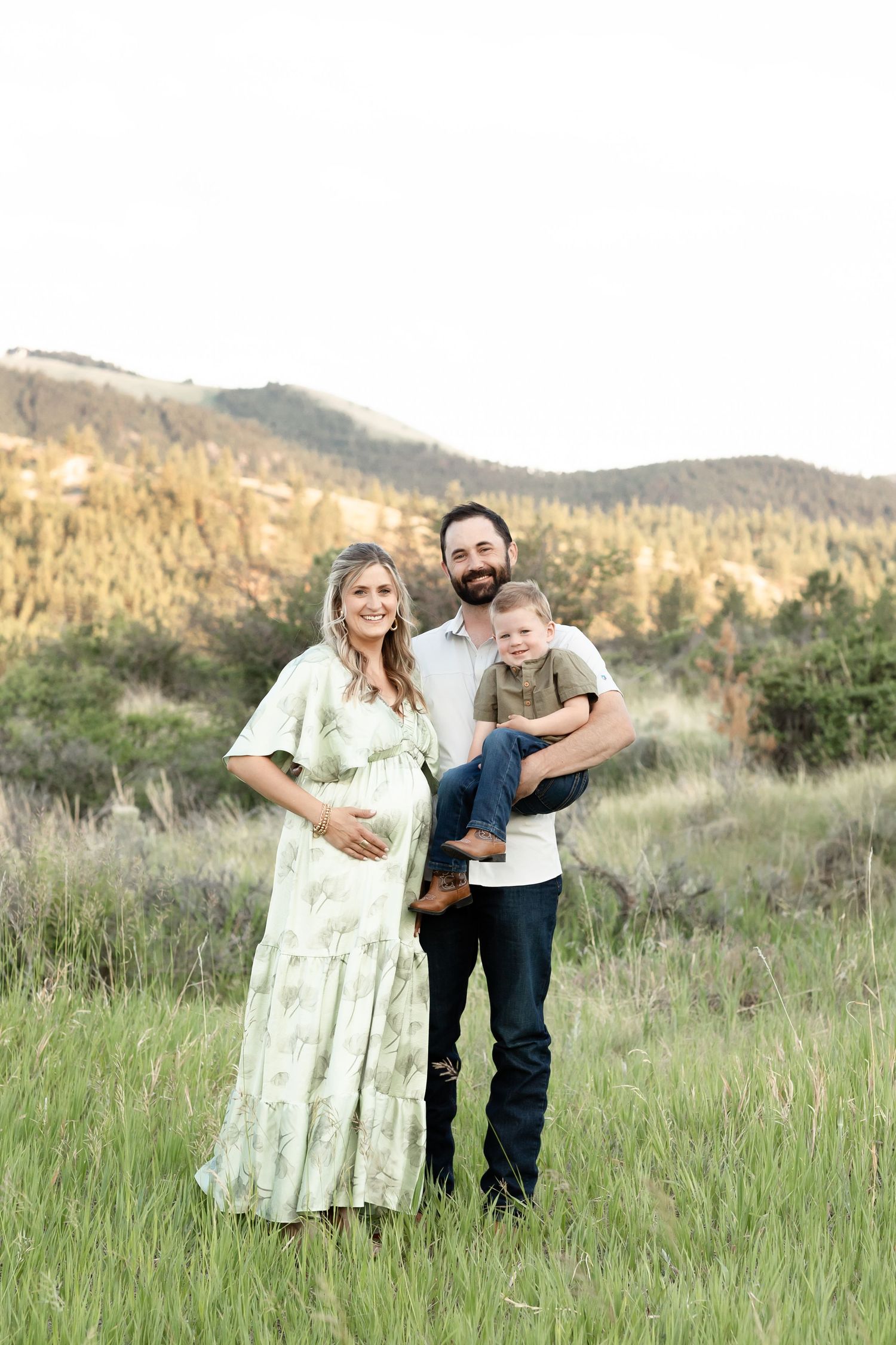 Smiling pregnant couple holding toddler son in green meadow with mountain backdrop.