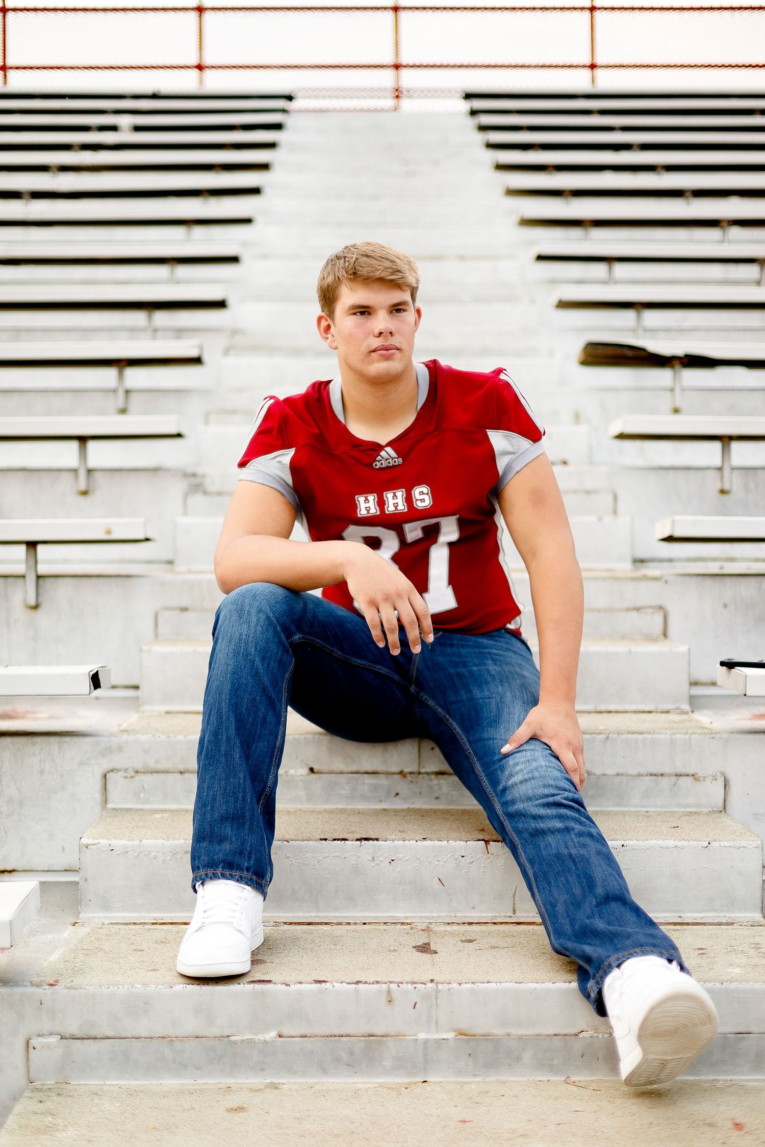 Teen in HHS #87 red football jersey sitting casually on bleachers wearing jeans and white sneakers.