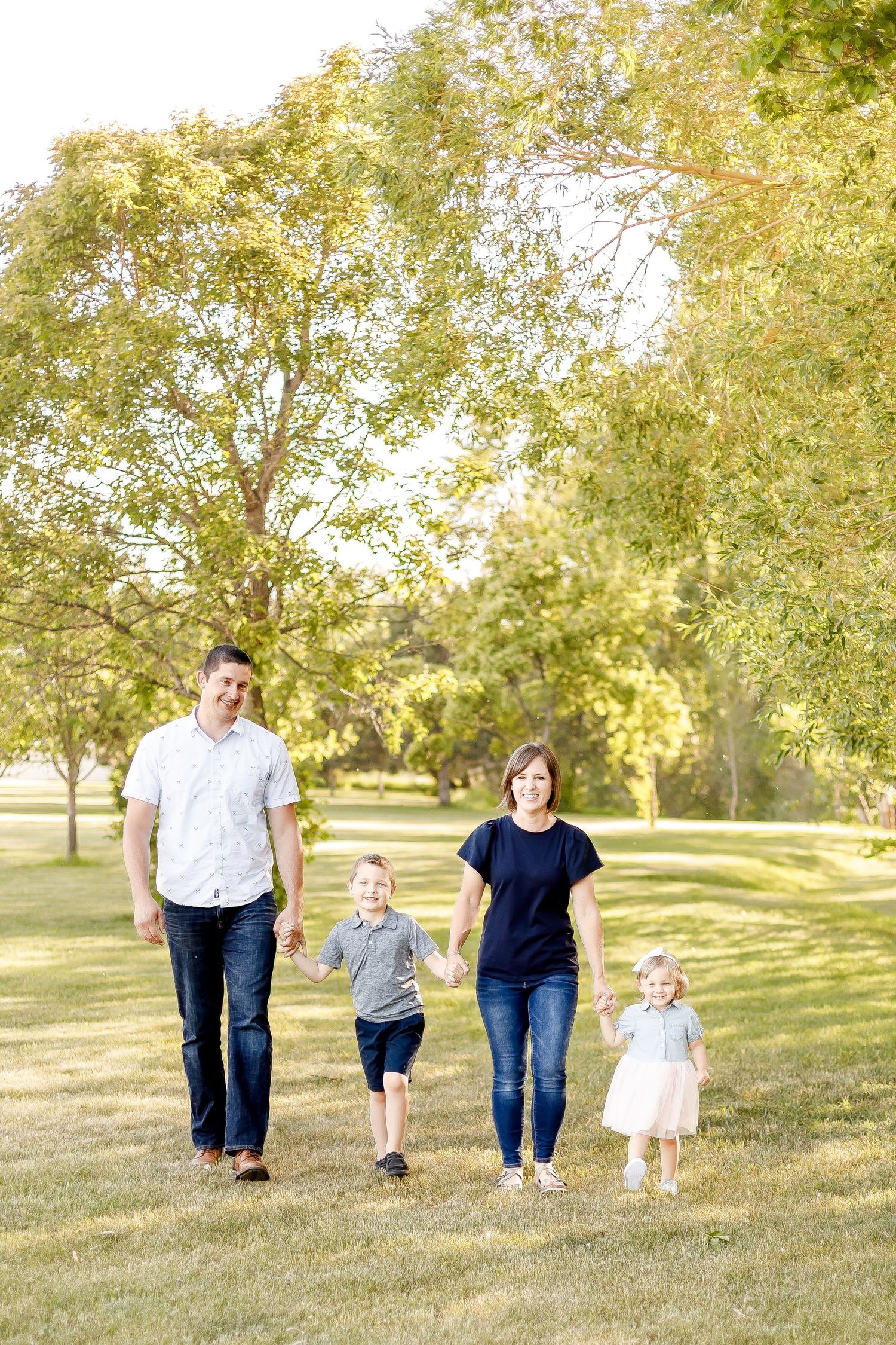 Happy family of four walks hand-in-hand through a sunny green park.