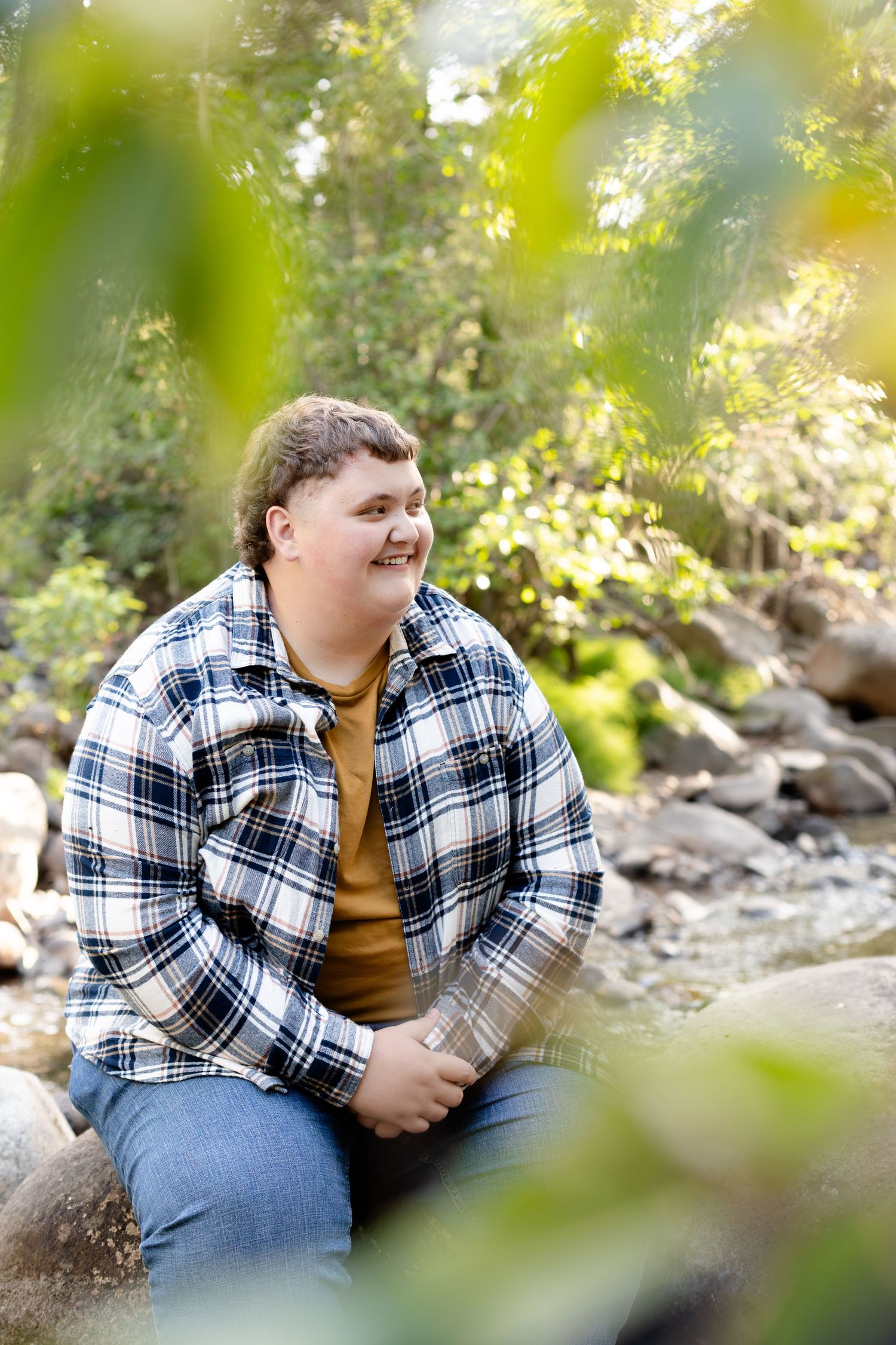 Young person in plaid flannel shirt sitting on rocks near a creek, smiling in a lush green forest.