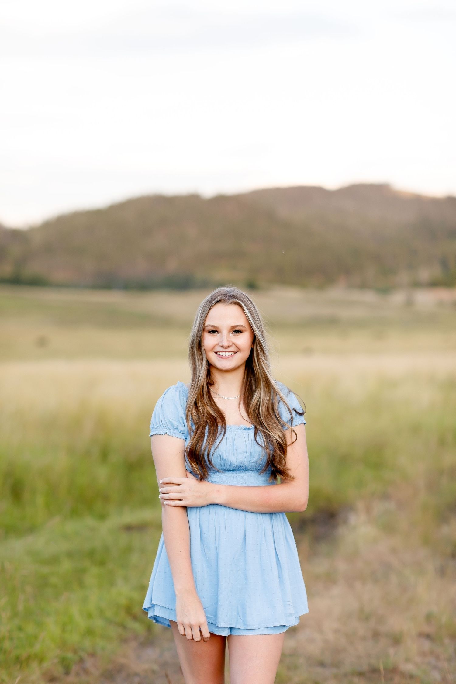 Smiling teenage girl in a blue dress standing in a golden meadow with mountains behind her.